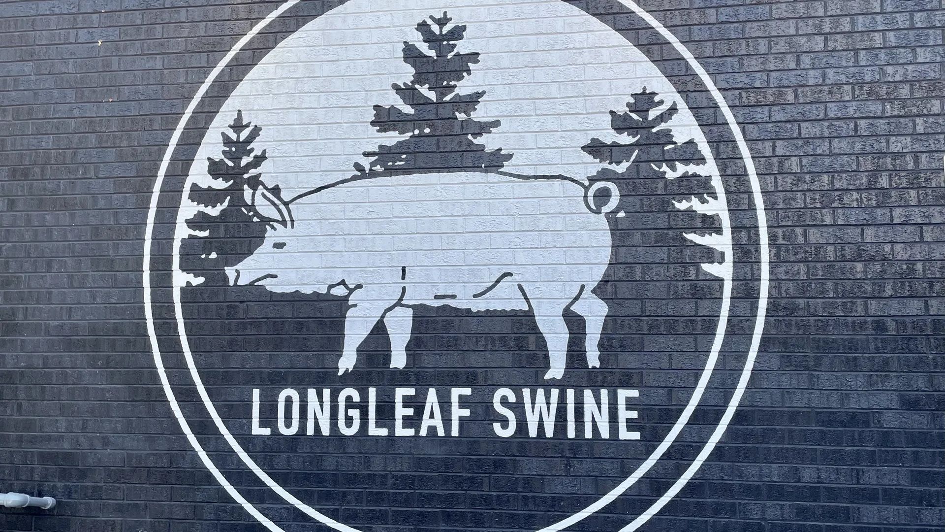 Black and white mural on a brick wall showing a pig silhouette with pine trees inside a circle, with the text "LONGLEAF SWINE" below the pig.
