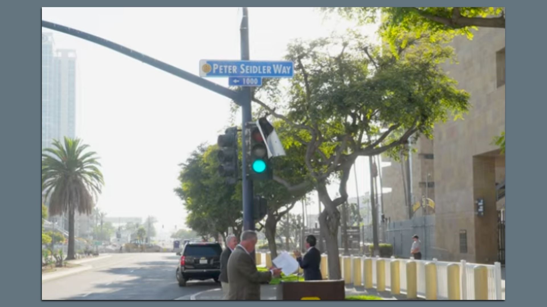 Street scene at Peter Seidler Way with green traffic light, trees, a few men in suits holding papers, a black SUV, and buildings in the background on a sunny day.