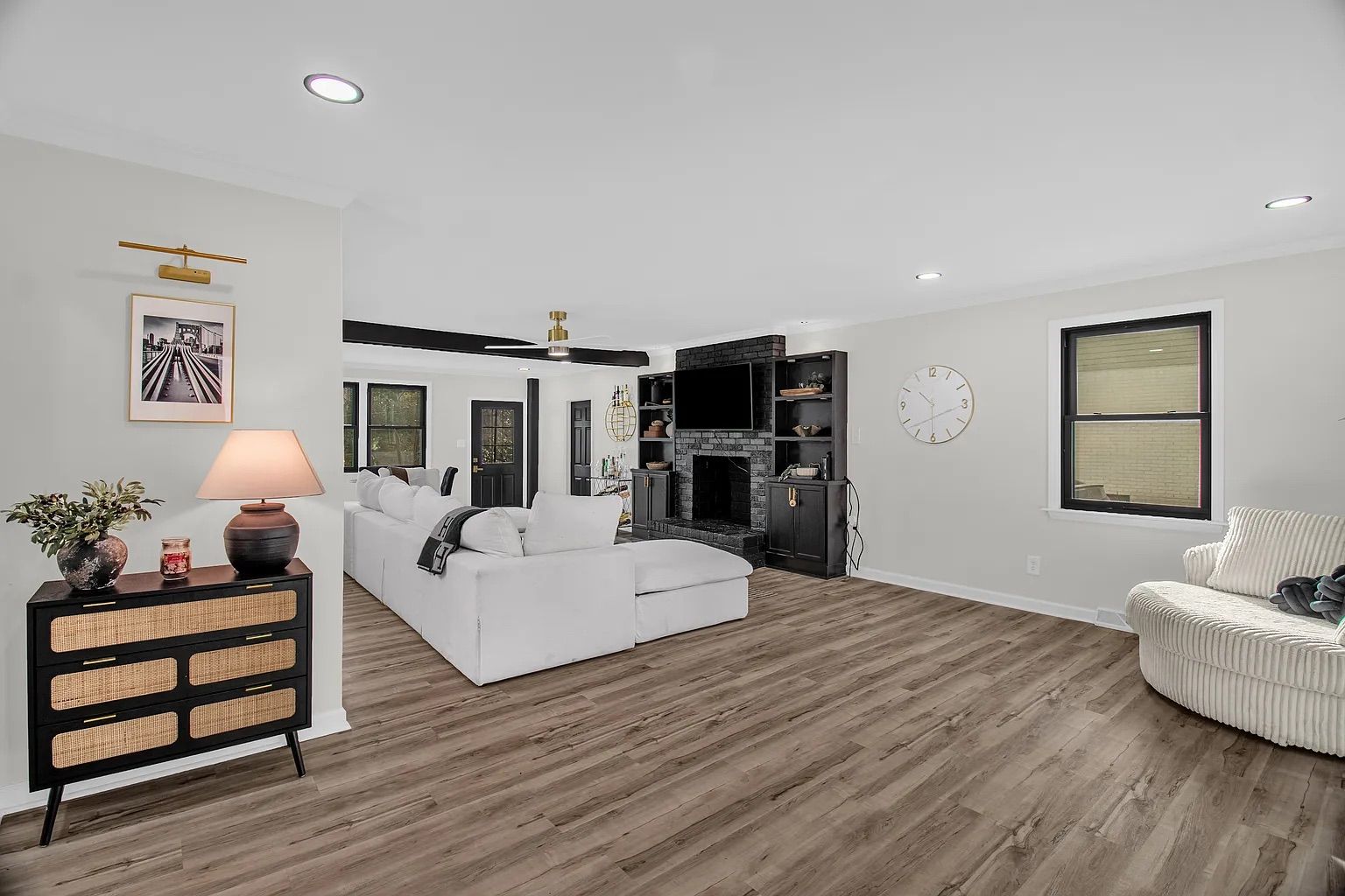 Modern living room with white sectional sofa, black brick fireplace with TV, wooden floor, white walls, black cabinets, round beige cushioned chair, black console table with lamp and plant.