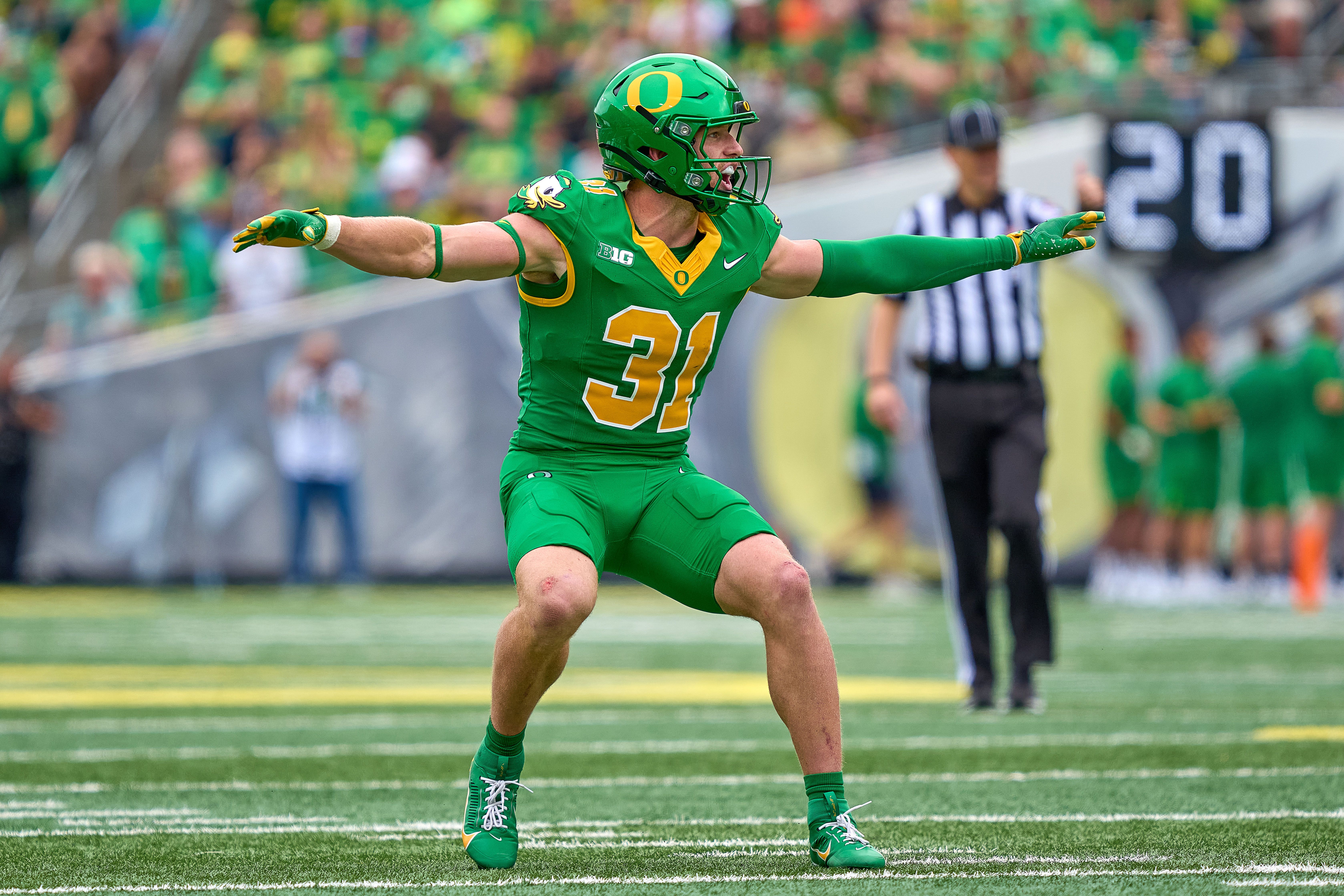 An Oregon Ducks football player in green and yellow with #31 stands on the midfield turf, arms extended in a wide stance. A green-clad crowd and a striped referee appear in the background.