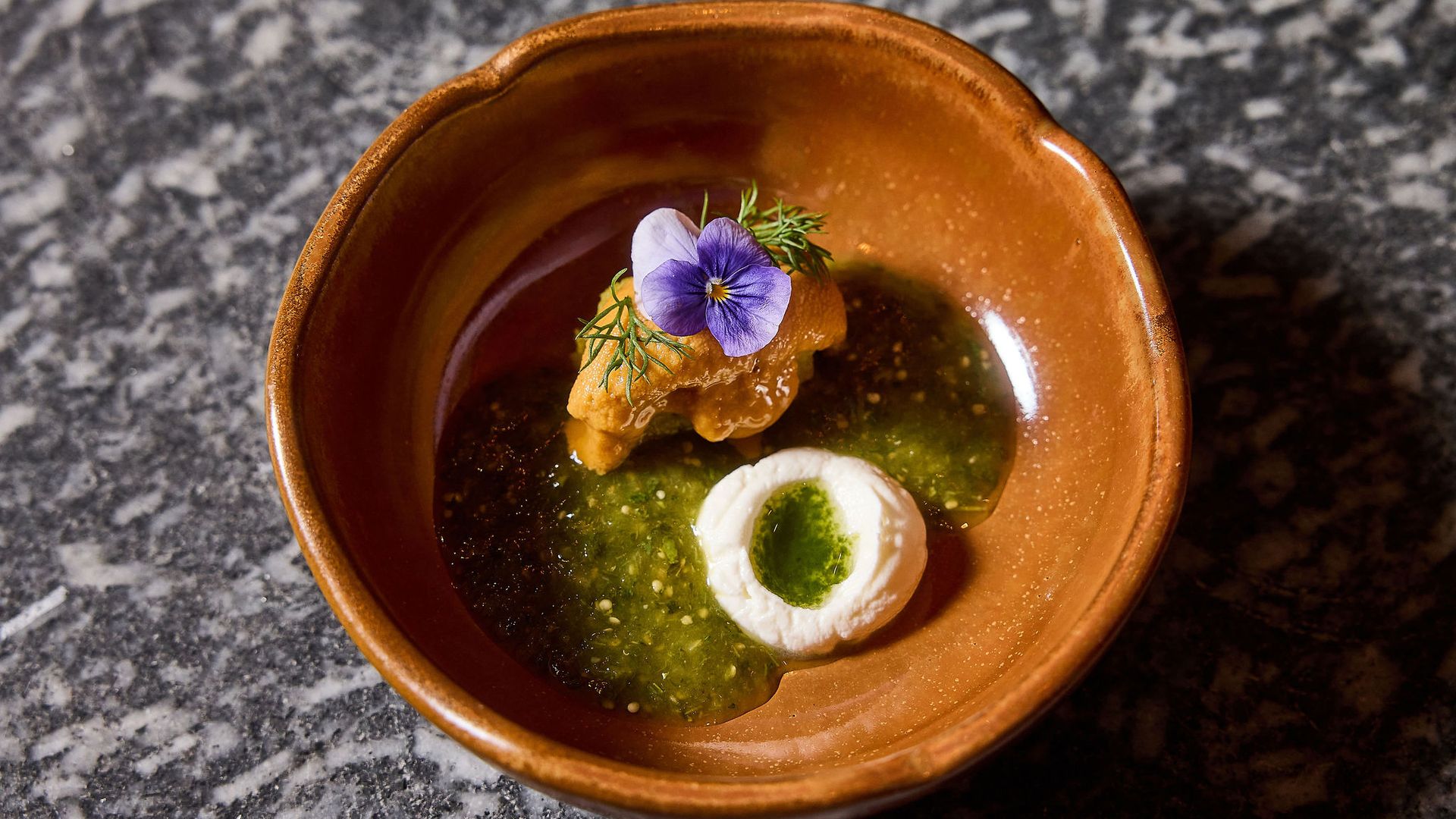 Top-down view of a brown ceramic bowl with green sauce and a fried piece topped by a purple pansy flower and dill, on a speckled granite countertop.