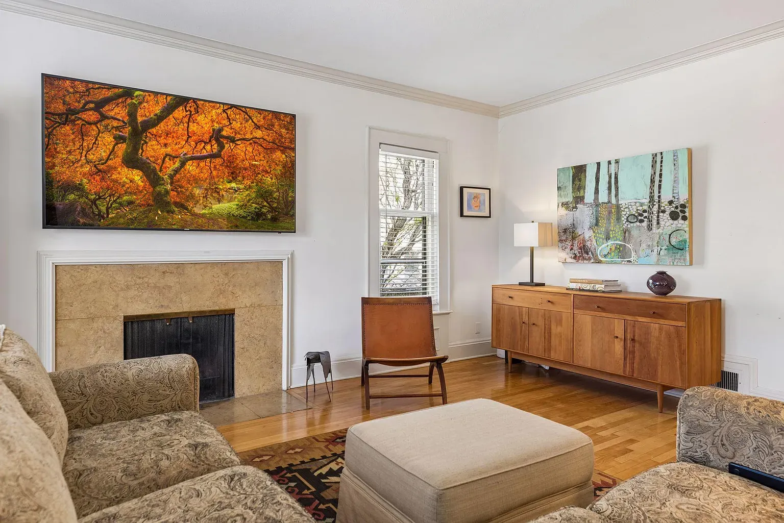 Bright living room with a fireplace and a large flat-screen TV above it. A beige patterned sofa and ottoman face a wooden sideboard with abstract art and a lamp; a brown chair sits by a window with blinds.