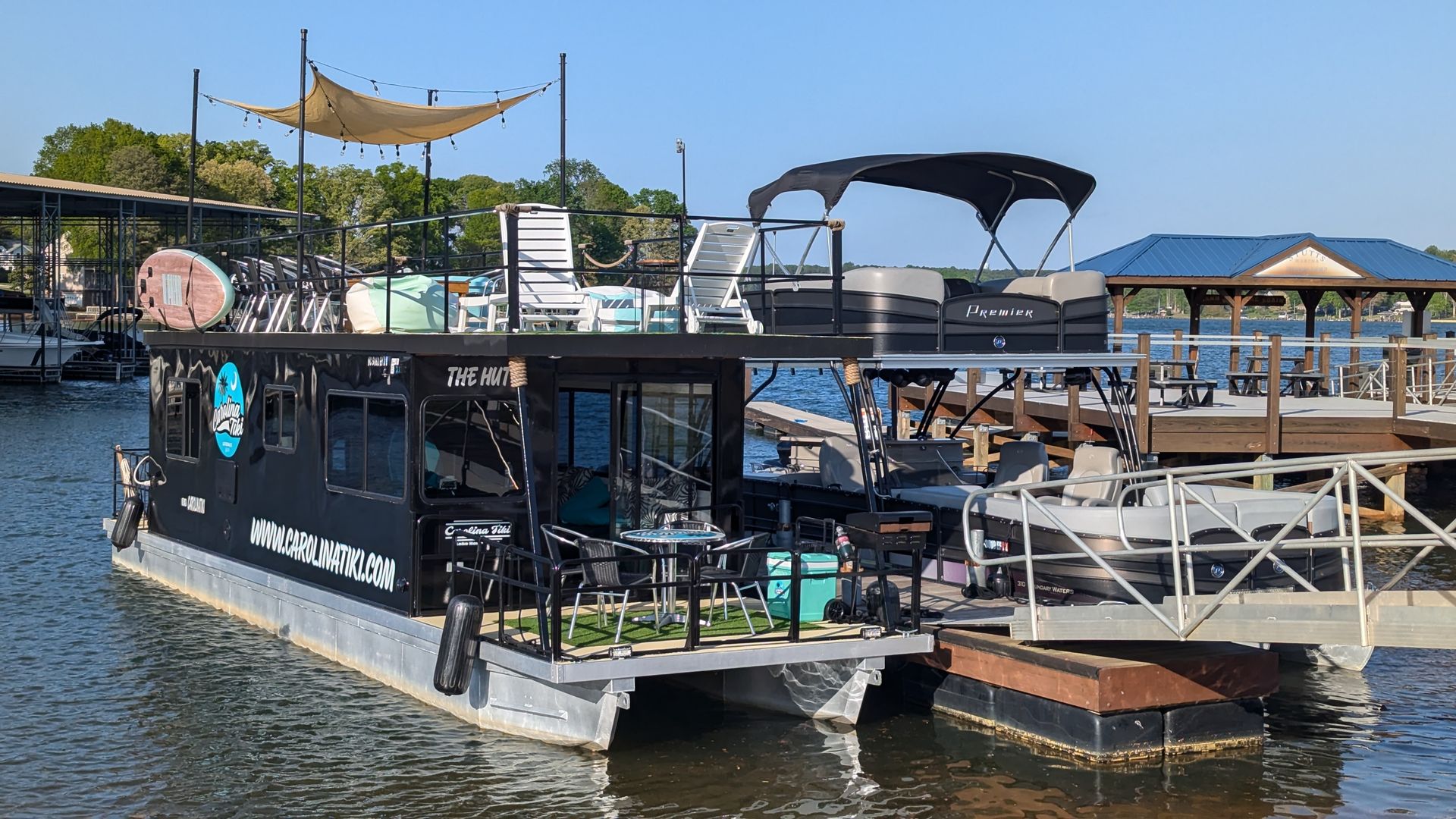 Sunlit marina with a black houseboat docked at a pier, tan canopy and string lights overhead, white lounge chairs on the deck, and a blue-roofed pavilion across calm water.