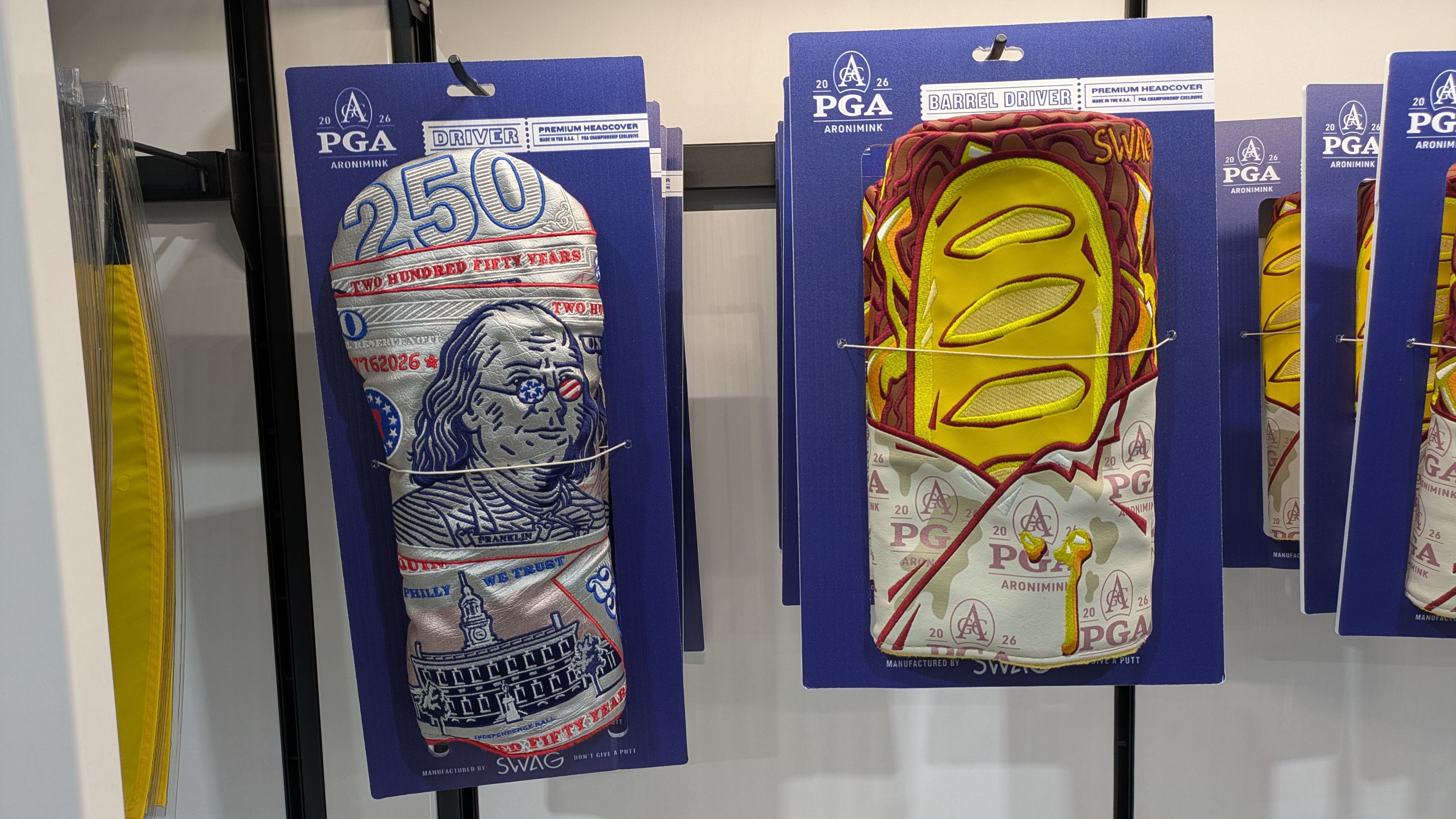Blue PGA display cards show two premium headcovers: left a silver 250-themed design with a vintage portrait; right a bright yellow and maroon barrel-style pattern with stitching.
