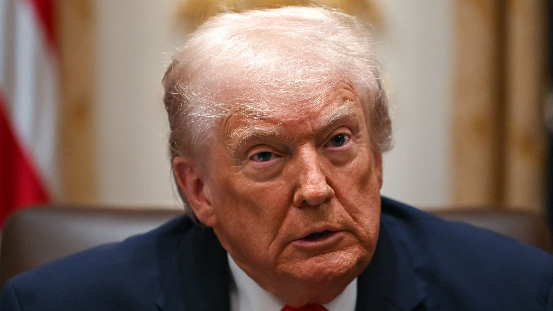 A gray-haired, wrinkly orange-fair skinned President Trump, wearing a navy jacket with a US flag pin at the top of his left lapel, white shirt and red tie, looks up from a brown leather chair in front of the red and white stripes of the US flag in the Cabinet Room of the White House. 