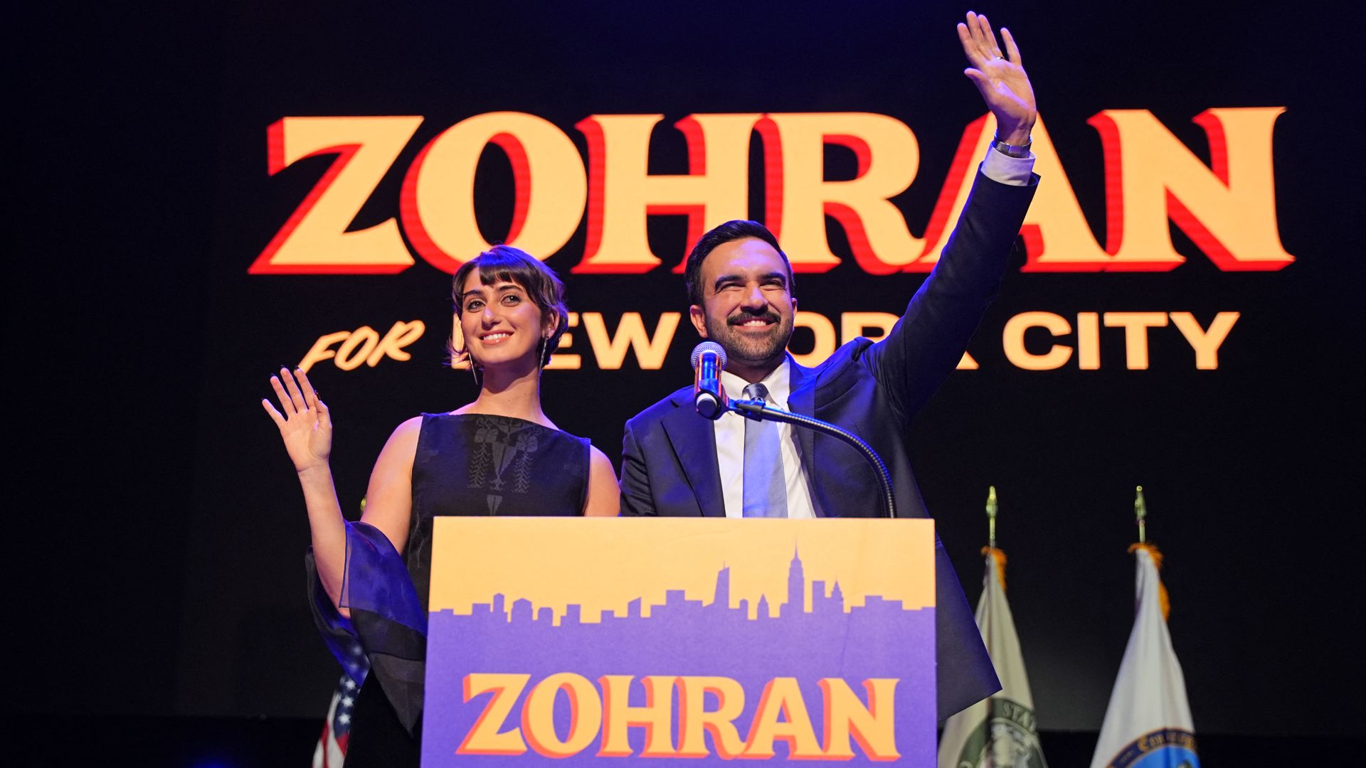 Rama Duwaji, wearing a black, sleeveless dress, waves with her right hand, and her bearded New York City Mayor-elect husband, Zohran Mamdani, wearing a navy jacket, white shirt and metallic blue tie, waves with his left before a podium bearing his name.