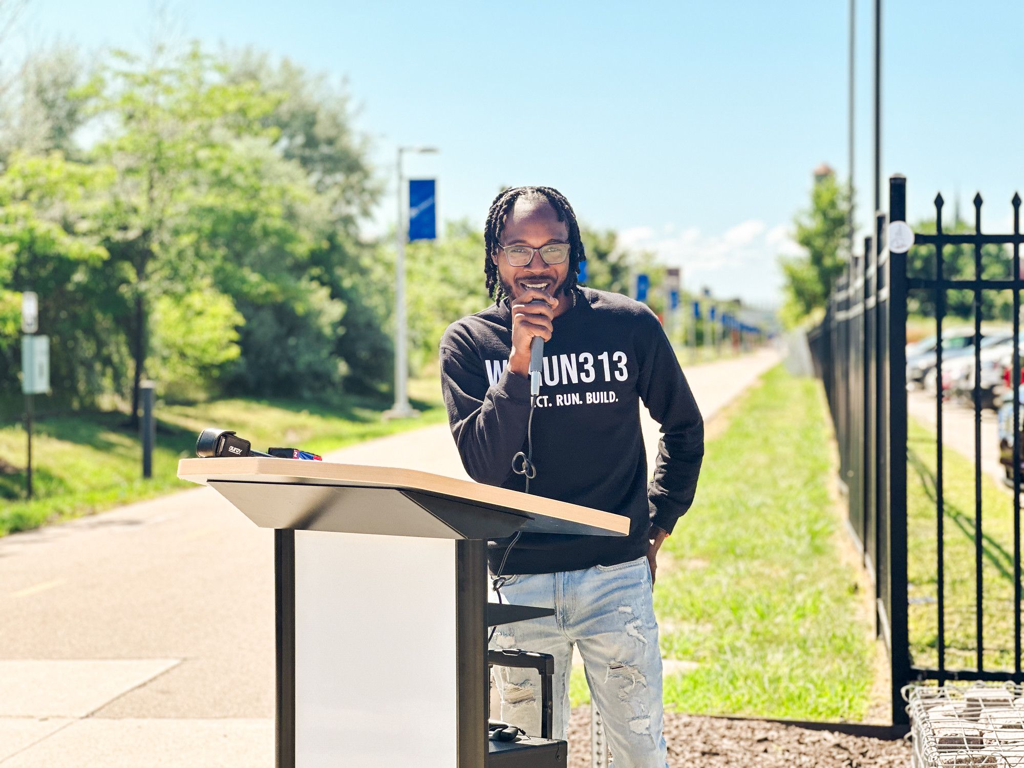 Lance Woods, We Run 313, standing in front of the Dequindre Cut, speaking at a podium in front of the pathway.