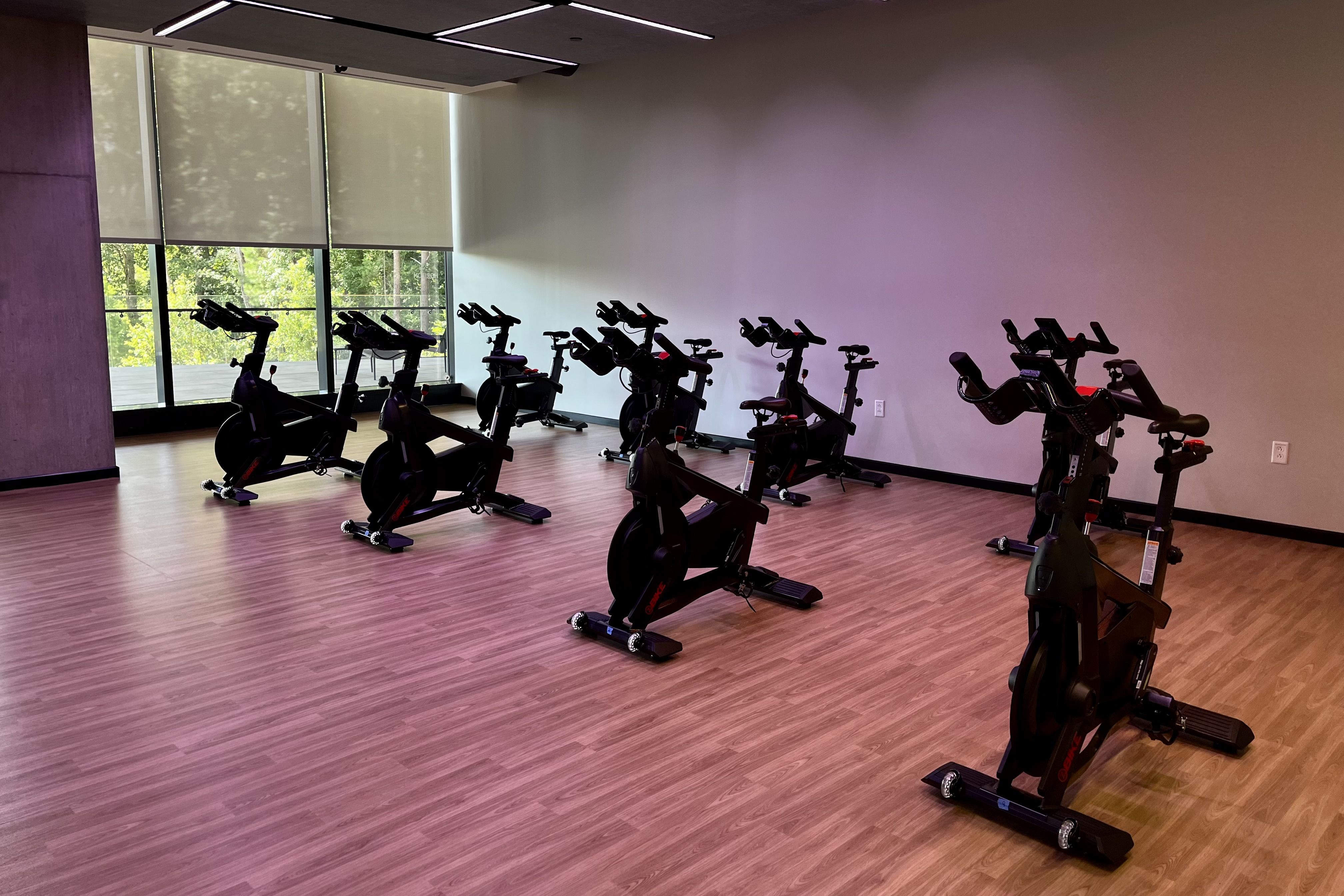 Empty indoor spinning bike studio with wooden floor, gray walls, large window with blinds partially closed, and eight black stationary bikes arranged facing the wall.