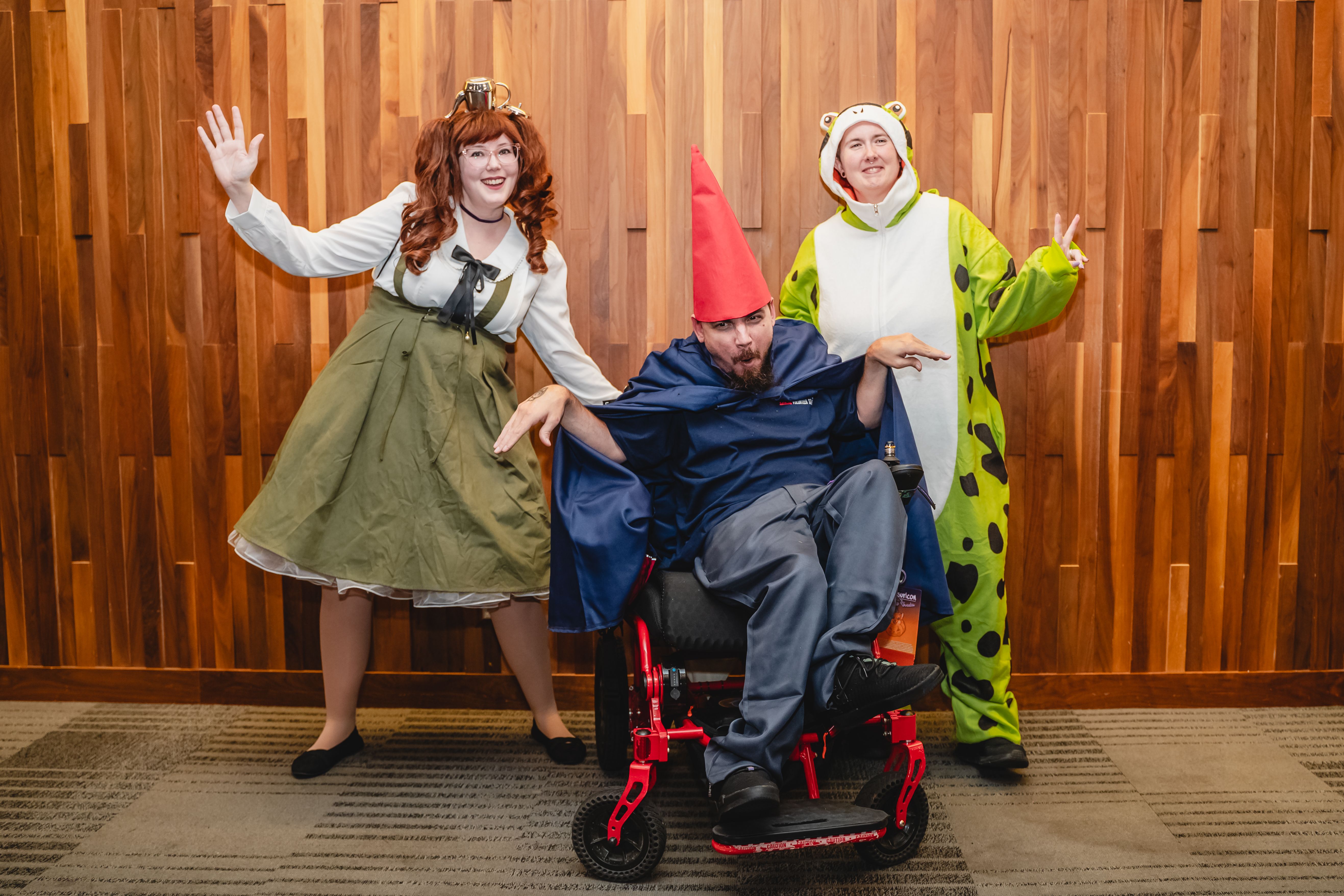 Three people in costumes posing against a wooden wall: one in an olive dress with a teapot hat, one in a green spotted frog onesie, and one in a blue cloak with a red cone hat in a red wheelchair.