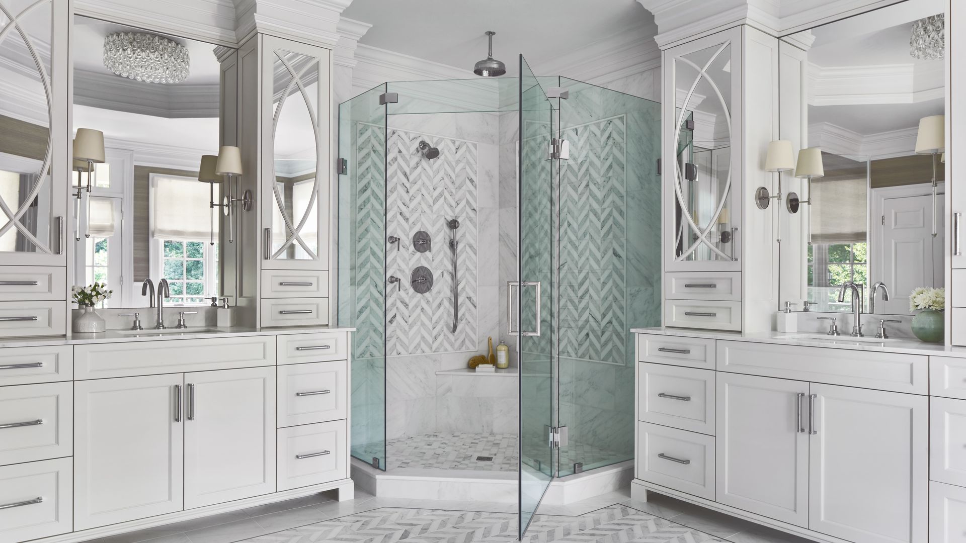 Luxurious white bathroom with dual vanities, glass-paned shower, and chevron marble floors. White cabinetry, marble countertops, and round mirrors create an elegant, bright space.