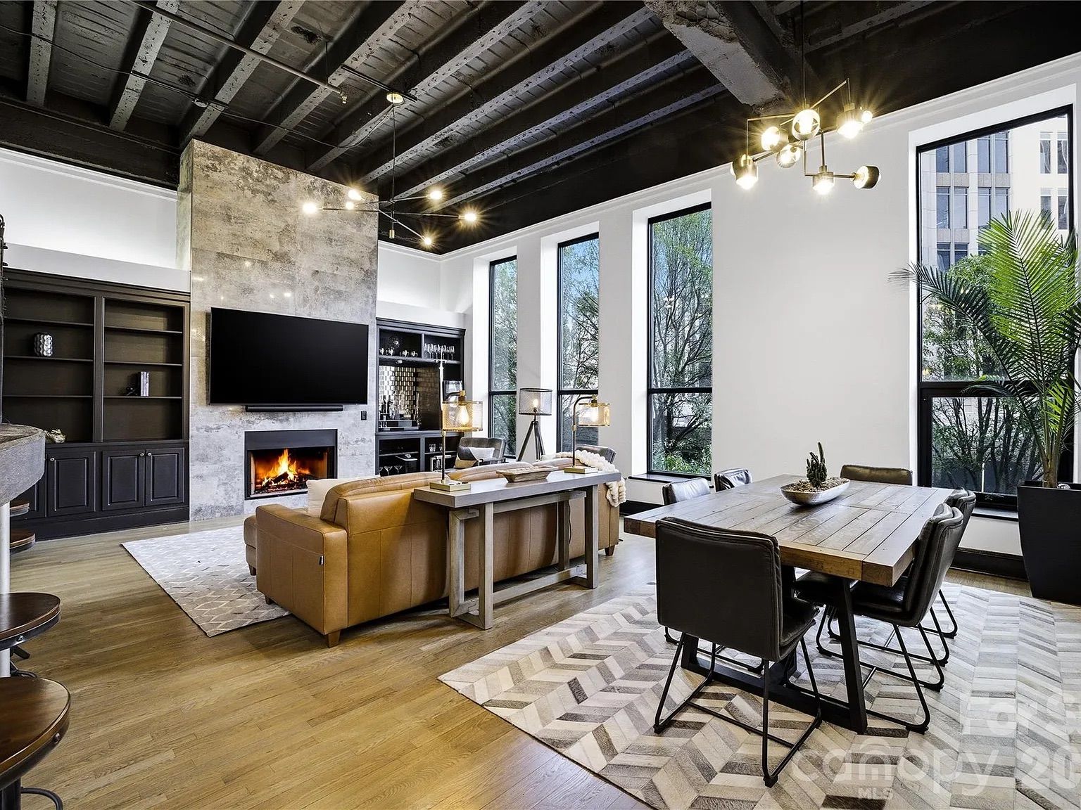 Modern living room with tan leather sofa, stone fireplace, large flat-screen TV, dark built-in shelves, wooden dining table with black chairs, and large tall windows letting in natural light.