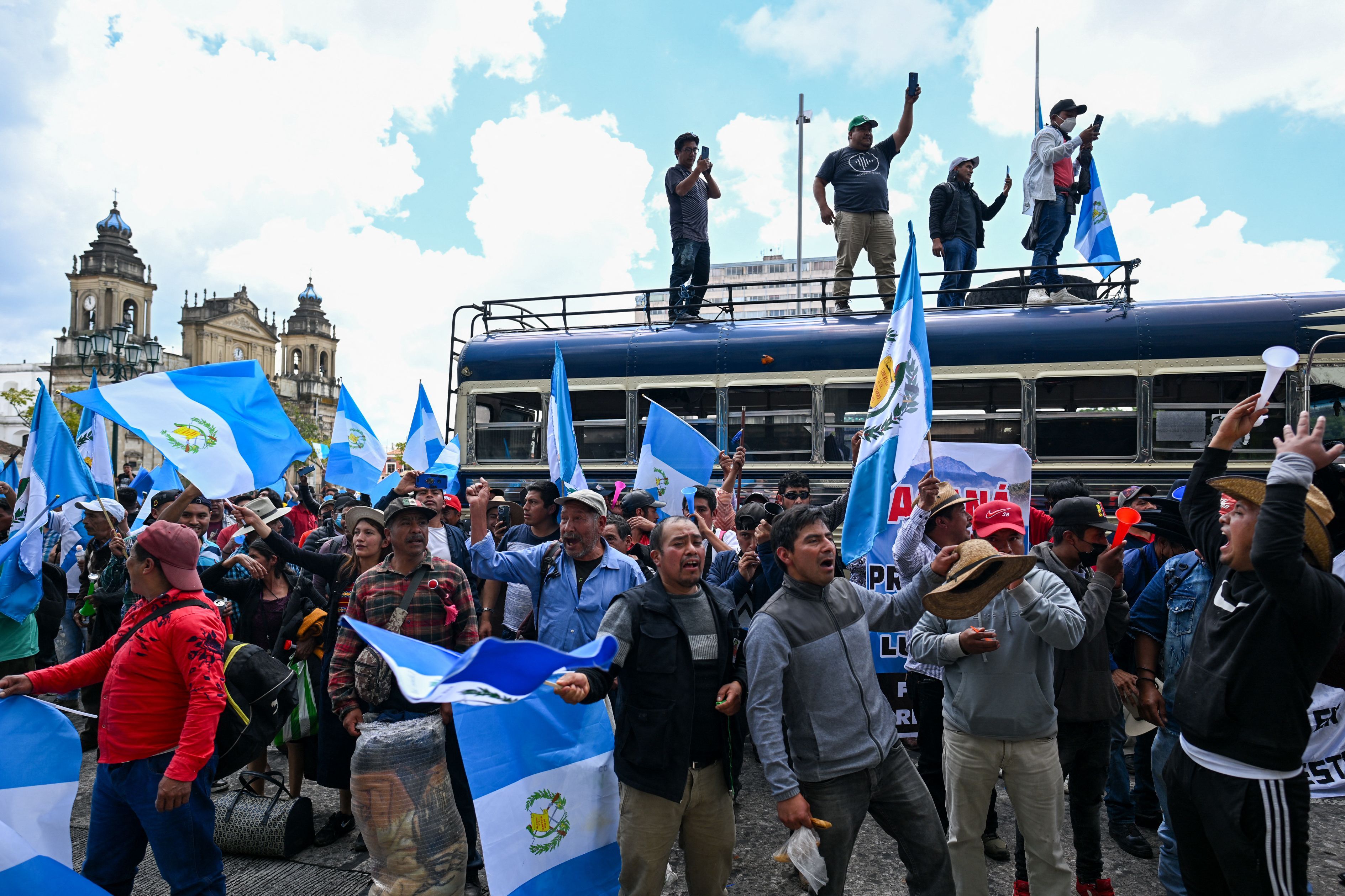 many protesters wave the blue and white Guatemalan flag while outside government buildings. some protesters are atop a bus. 