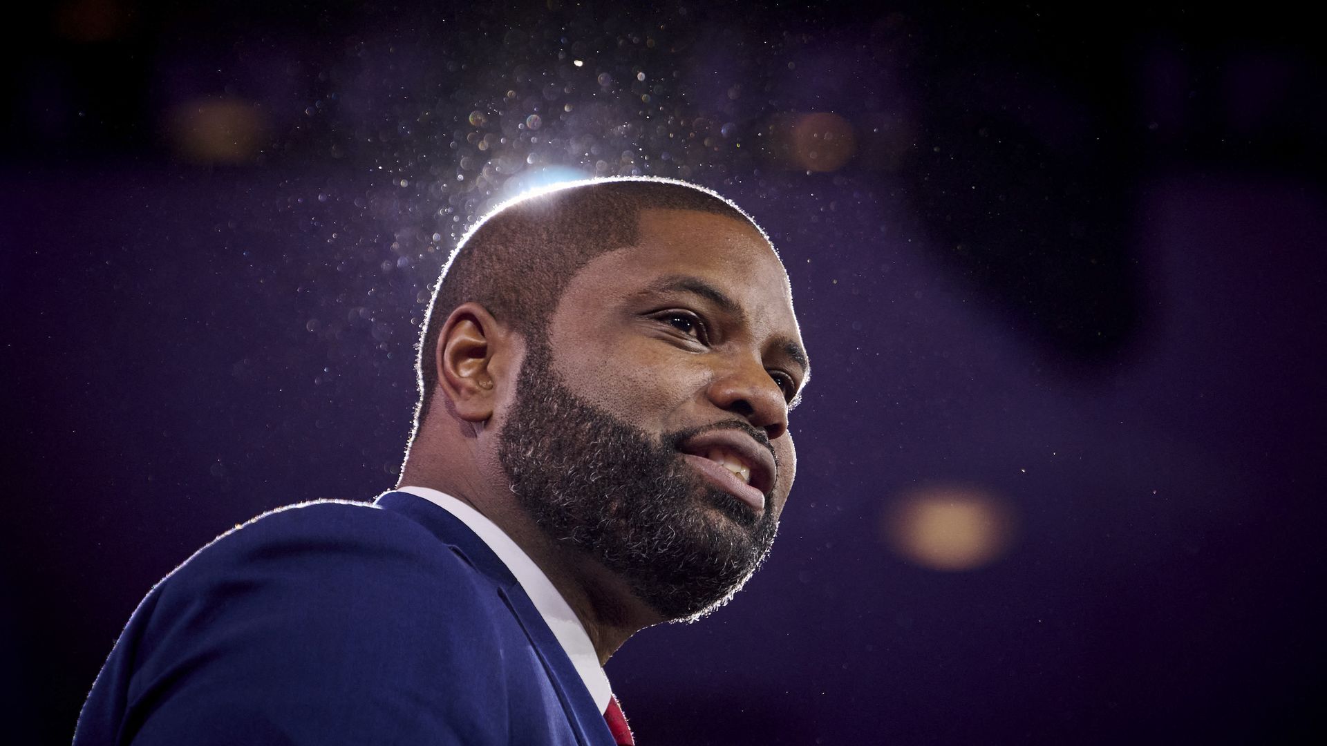 A man in a navy suit and red tie looks to the right with light spilling out from behind his head.