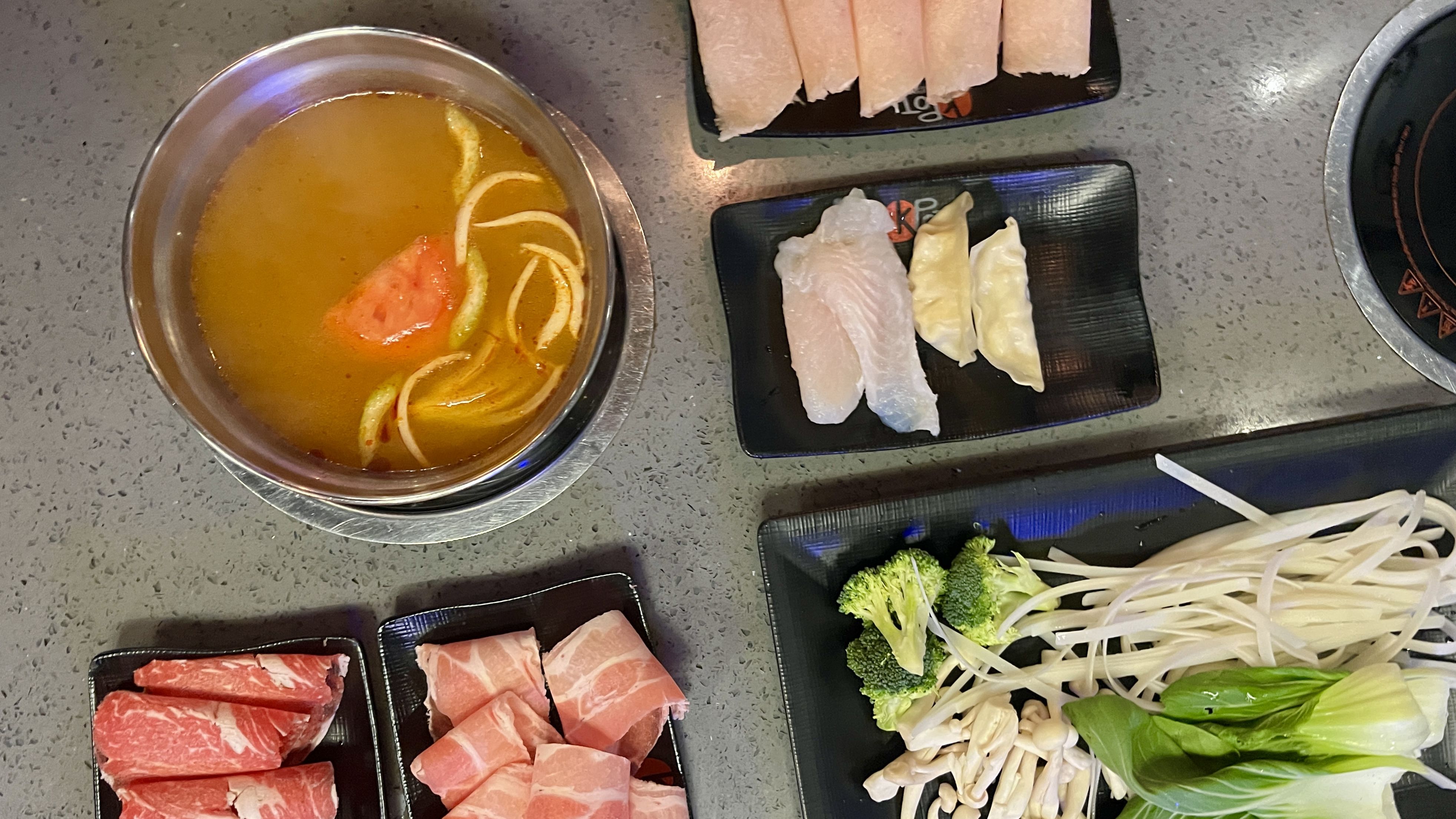 Top-down view of hot pot meal at KPOT in Overland Park with a bowl of broth with vegetables, plates of raw thinly sliced meat, dumplings, fish slices, broccoli, mushrooms, and leafy greens on a gray countertop.