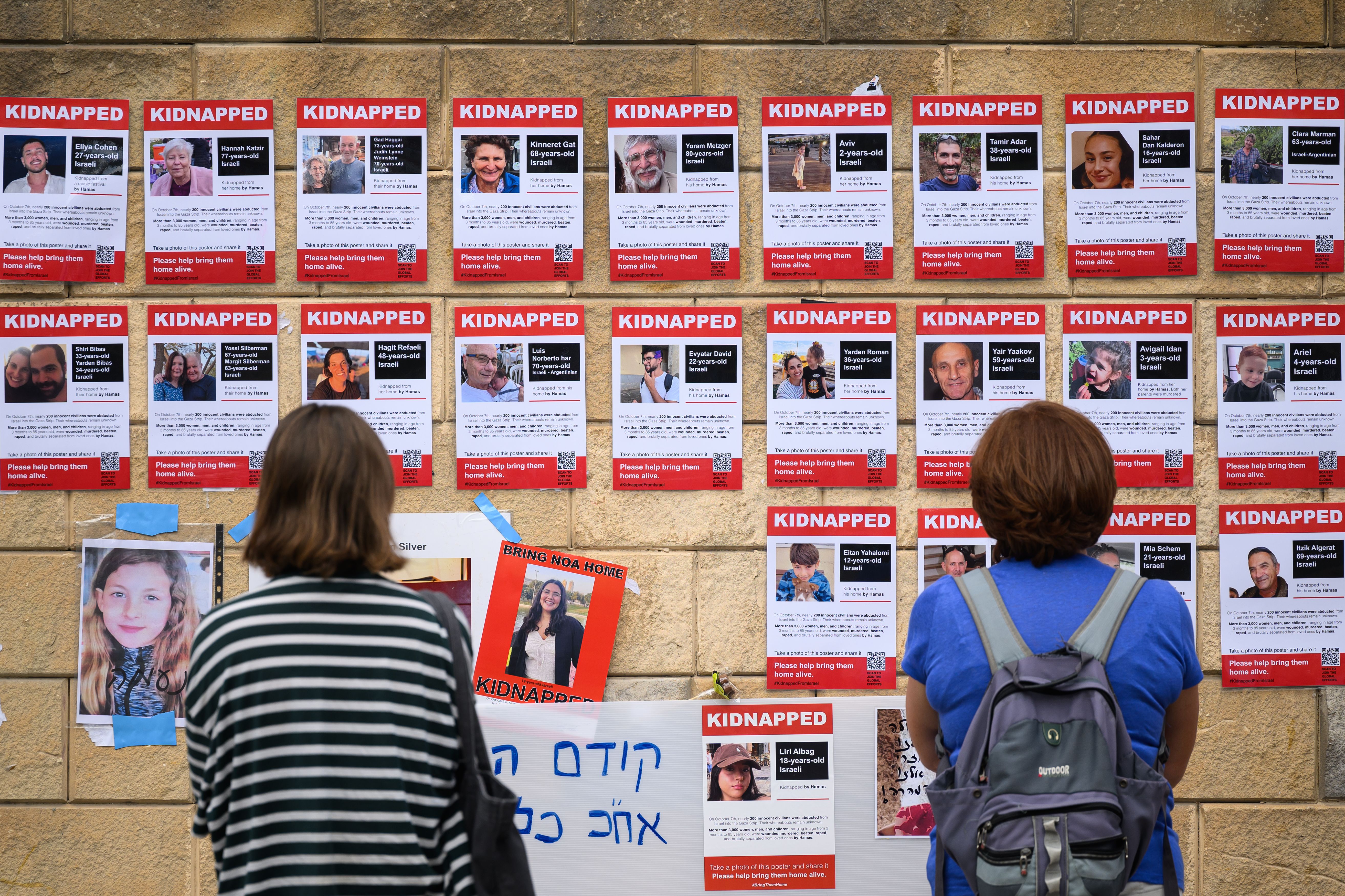 Two people looking up at a wall covered in posters showing faces of hostages with the word "KIDNAPPED" at the top of each poster. 