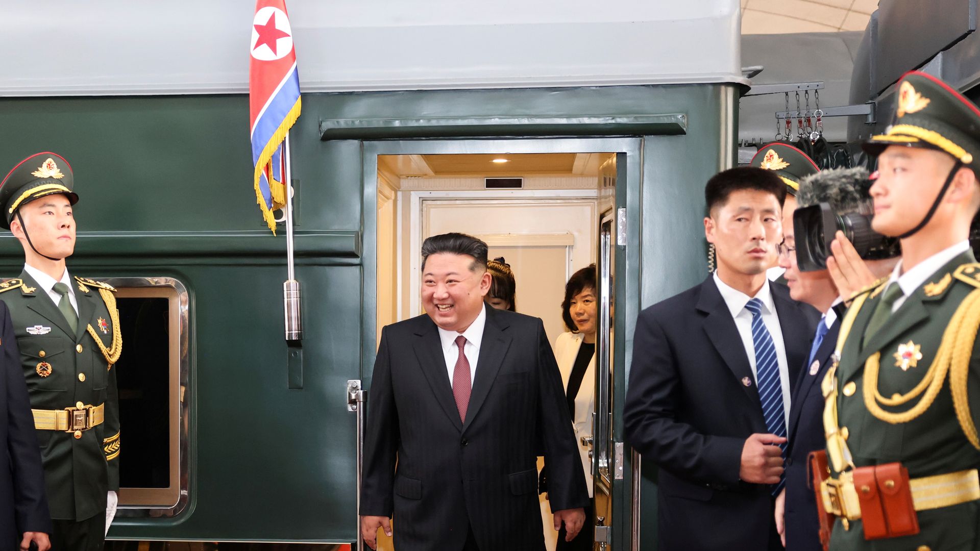 North Korean leader Kim Jong-un, wearing a black suit, white shirt and red tie, stepping off a green train carriage that's surrounded by North Korean military guards.