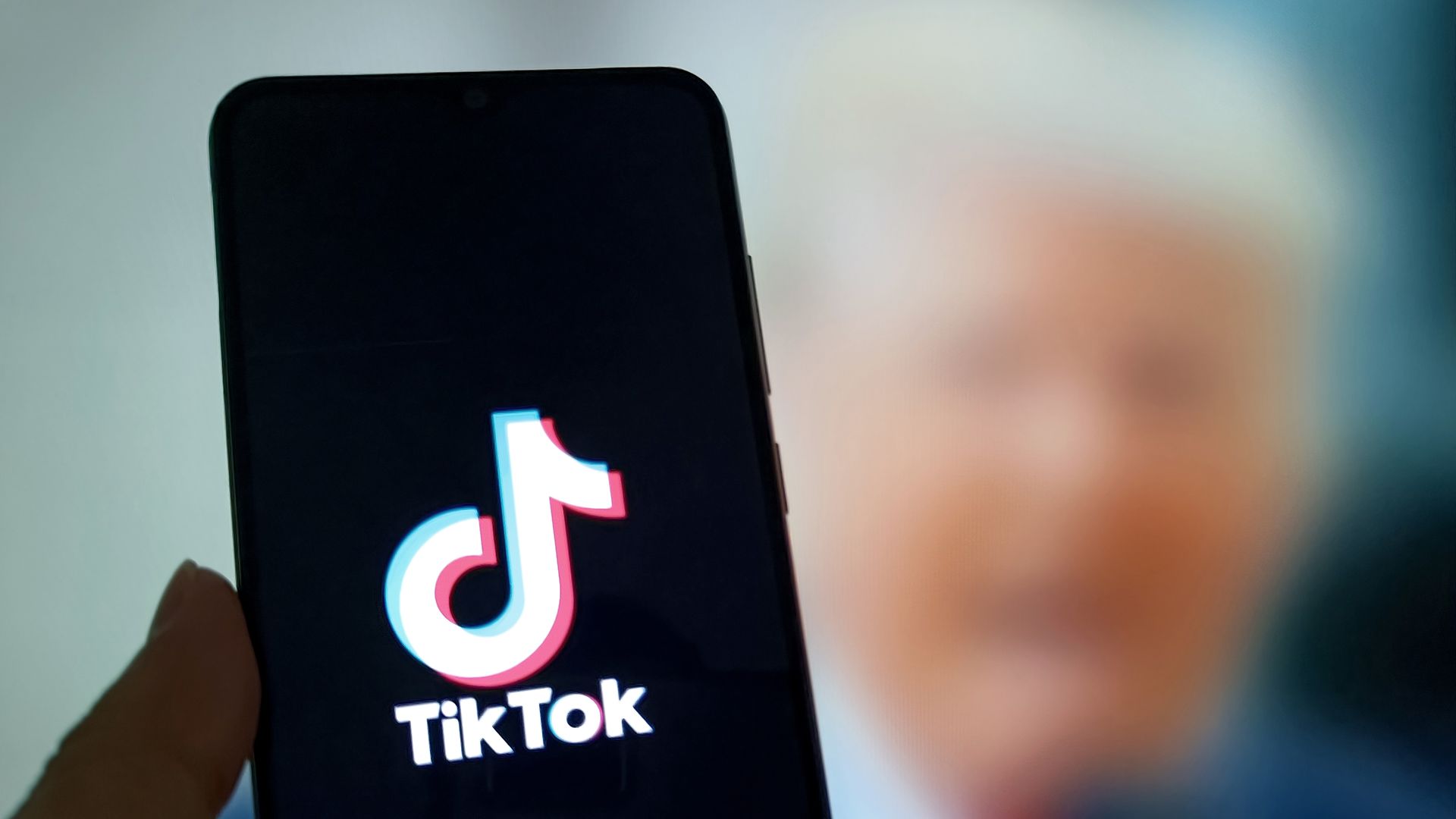 A hand holding a phone showing the TikTok logo, in front of a blurred picture of President Trump. 