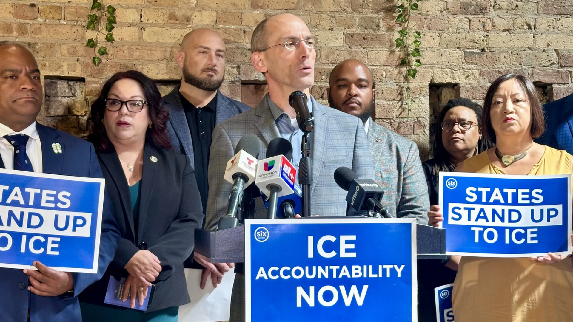 Speaker at a podium with microphones, flanked by diverse supporters; blue signs read "ICE ACCOUNTABILITY NOW" and "STATES STAND UP TO ICE" against a brick wall.