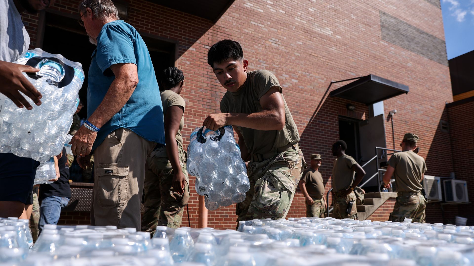 People, including service members in camouflage pants and olive shirts, unload and organize cases of bottled water outside a brick building under a clear sky.