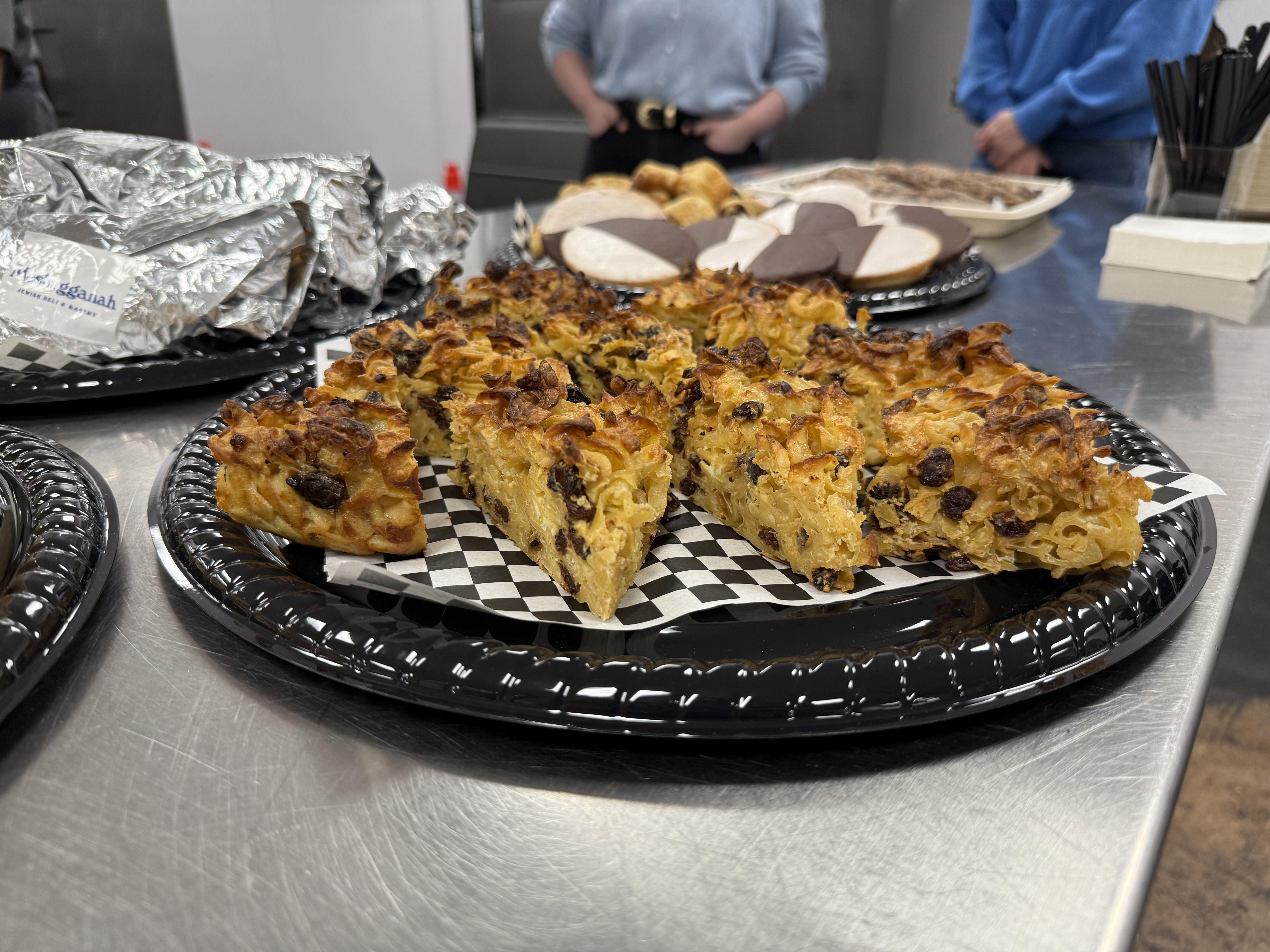 Close-up of triangular slices of baked dessert with raisins on black tray with checkered paper, surrounded by cookies and wrapped sandwiches on stainless steel table.