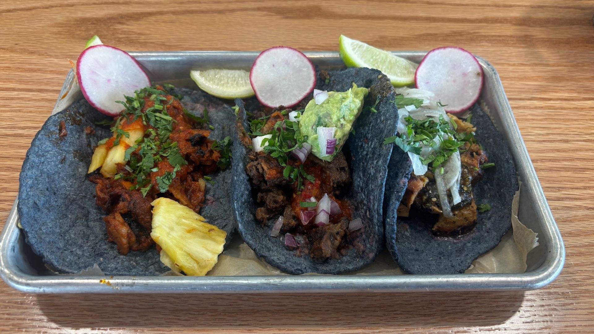 Three blue corn tacos on a metal tray with radish slices, lime wedges, cilantro, chopped onions, guacamole, pineapple chunks, and various meats and salsa toppings.