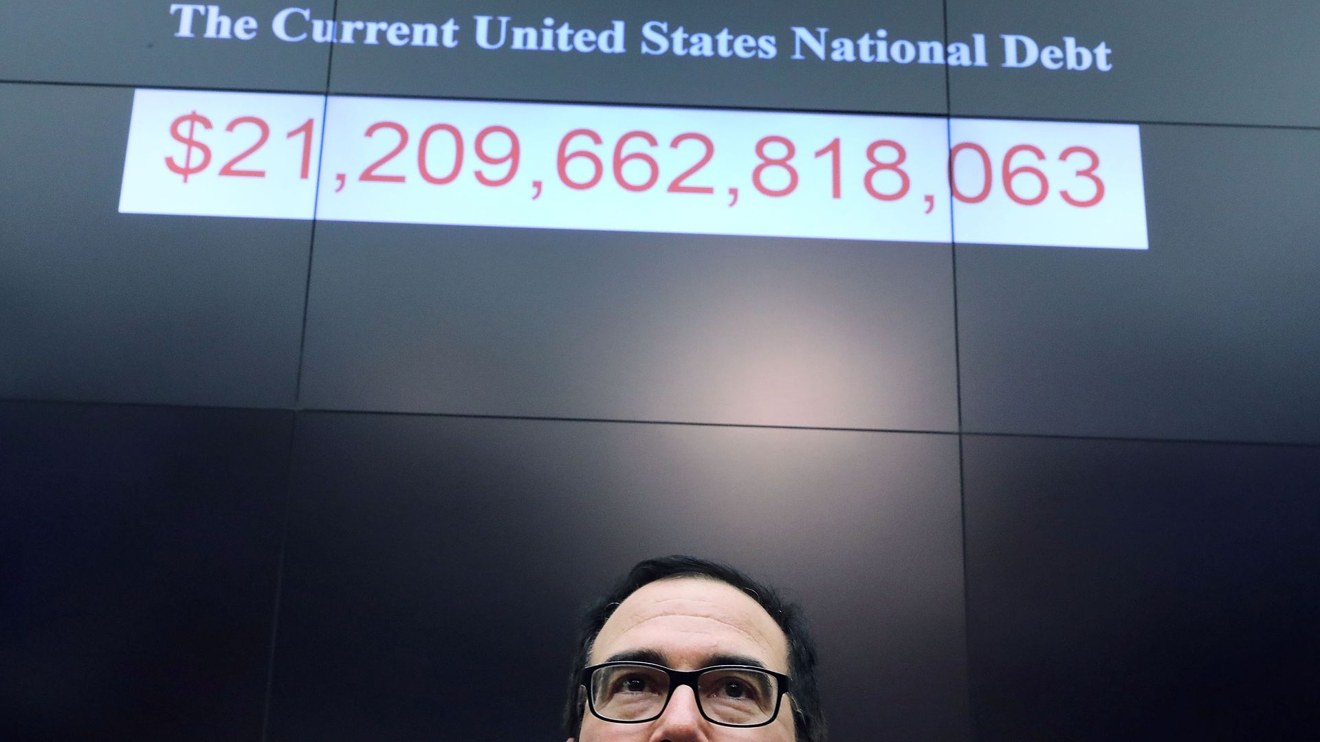 Steven Mnuchin standing under a sign that details the U.S. National Debt. 