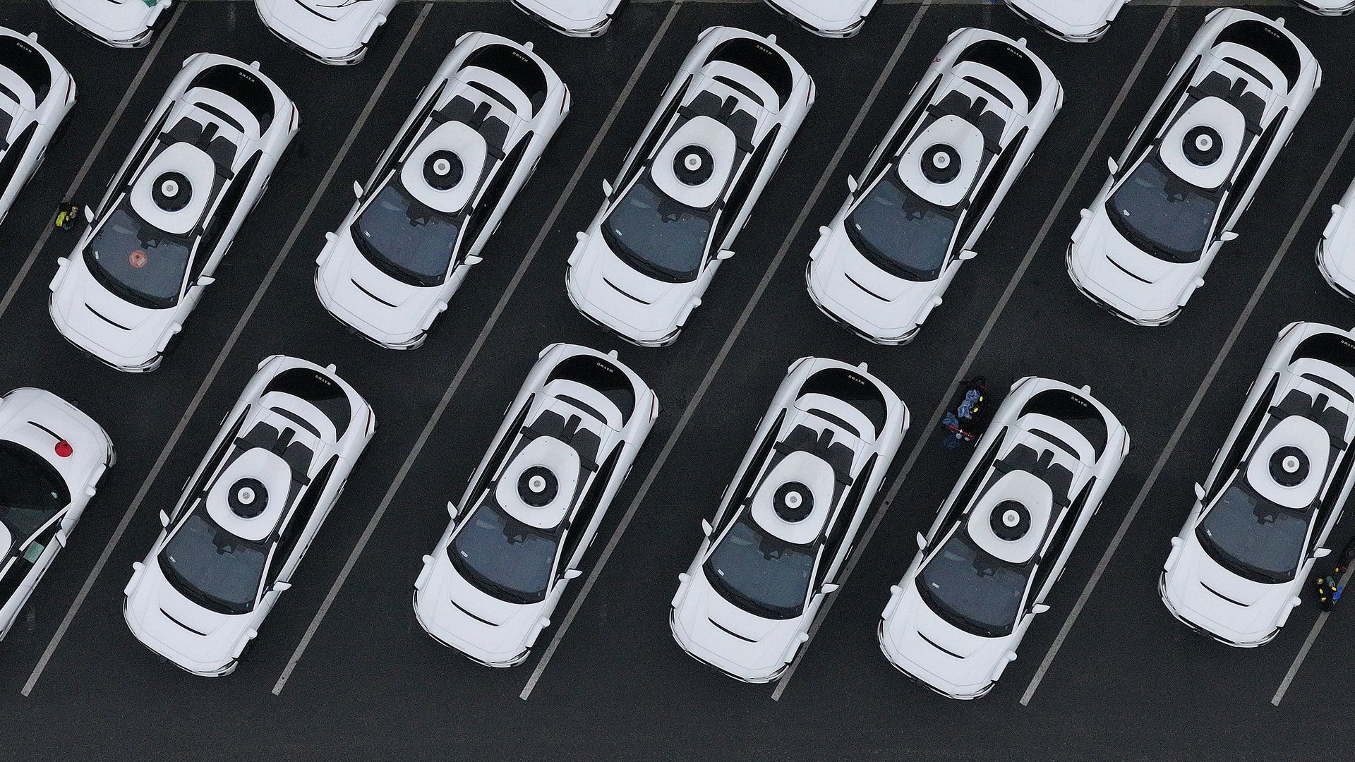 Aerial view of multiple white self-driving cars lined up diagonally in a parking lot with black roofs and circular sensors on top, separated by white parking lines on dark pavement.