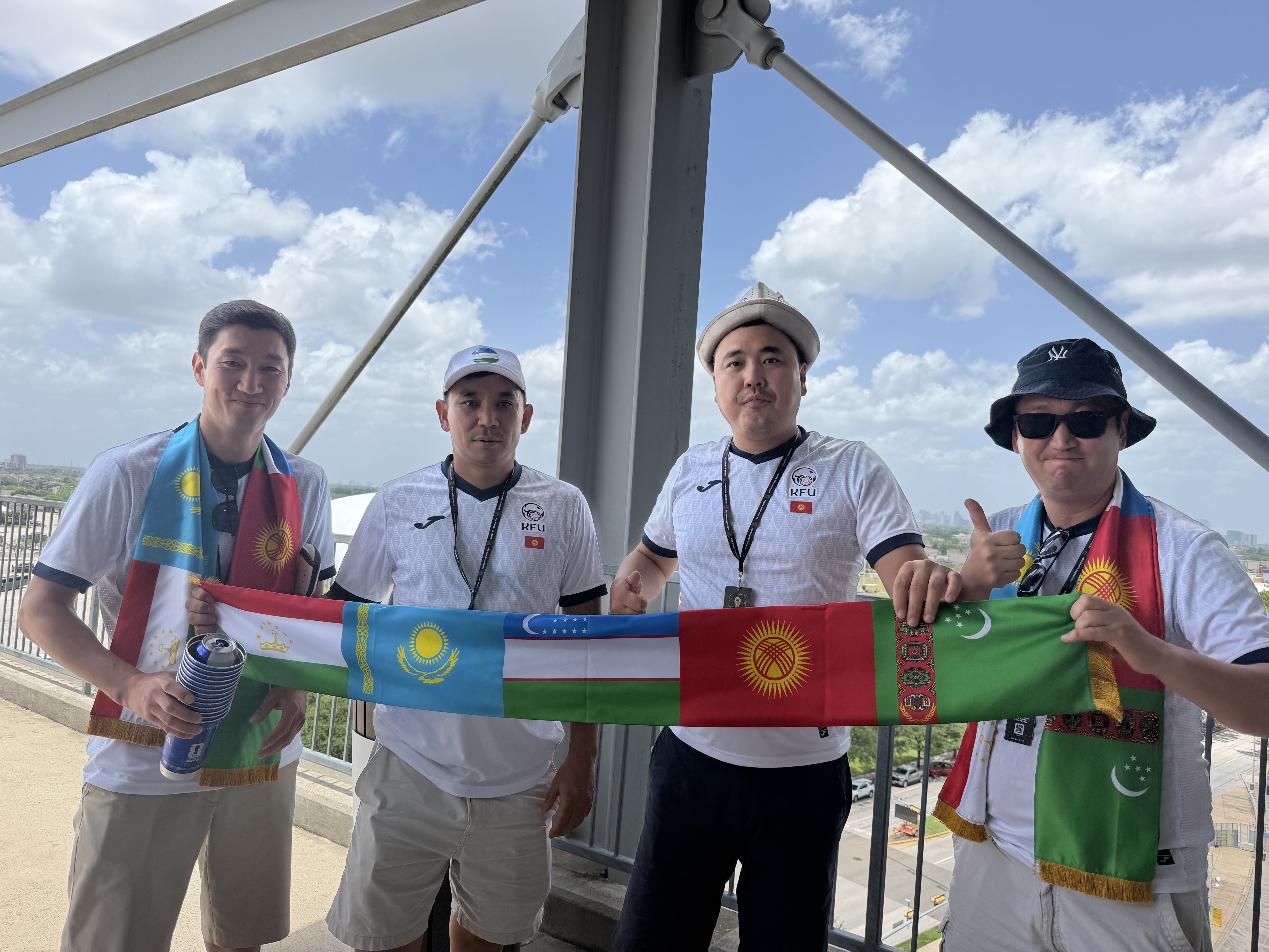 Four men in white jerseys stand on a metal balcony, holding a long banner of multiple national flags. The bright blue sky with clouds and steel beams looms behind.