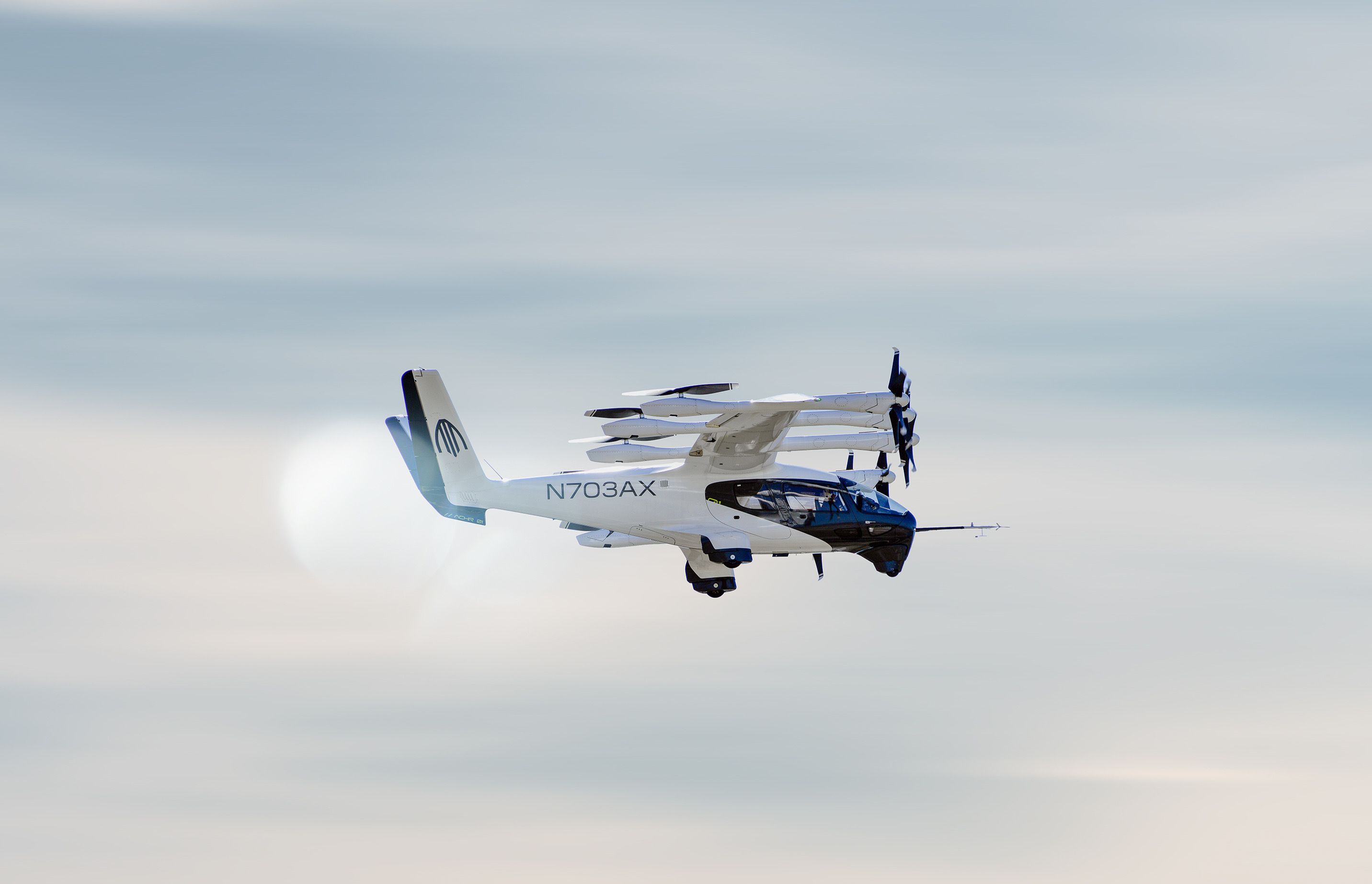 A white and blue tilt-rotor aircraft in midflight against a pale blue sky with wispy clouds; blades extended, registration N703AX on fuselage, logo on tail, landing gear retracted.