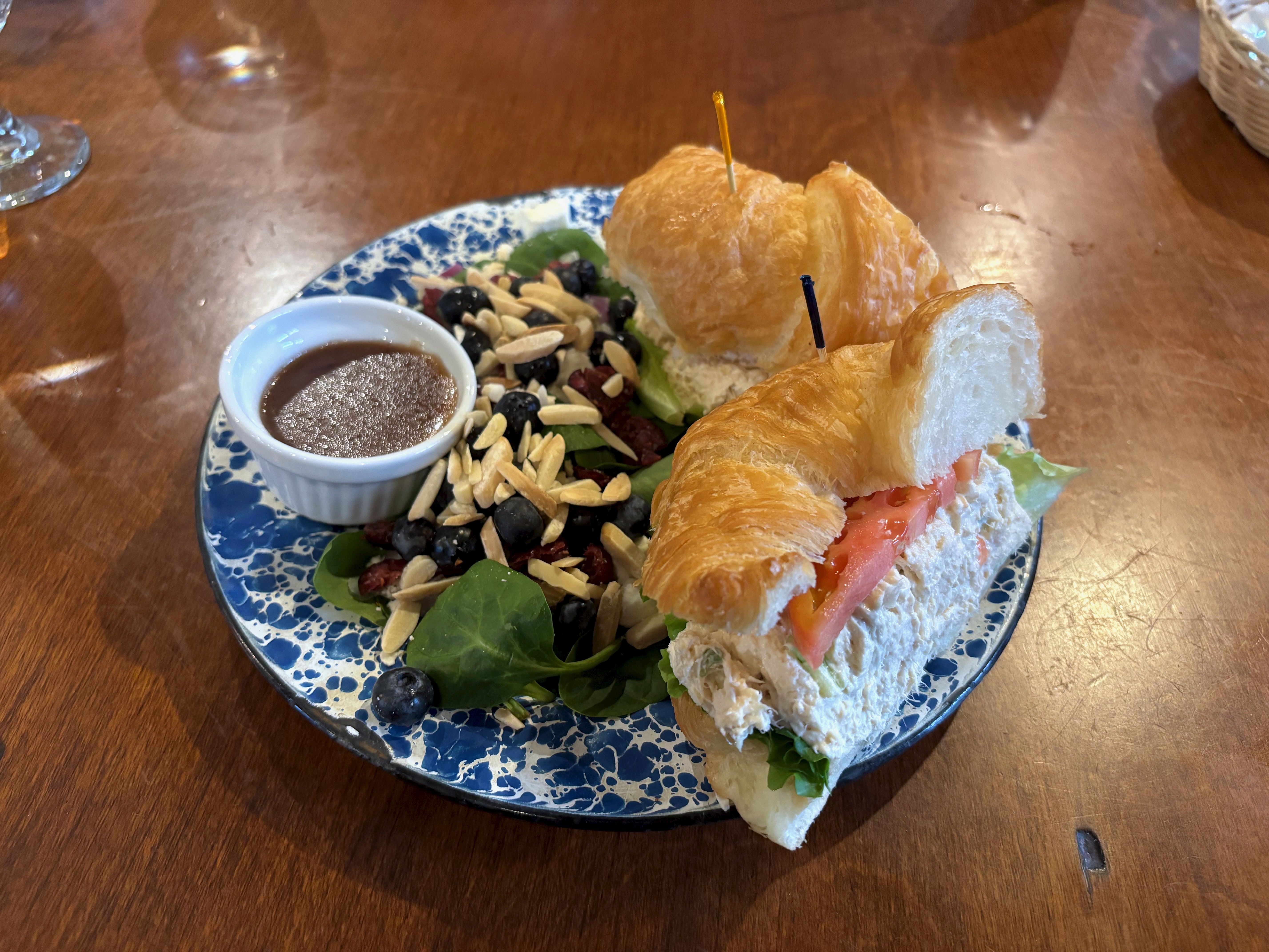 Plate with two croissant chicken salad sandwiches, salad with spinach, blueberries, slivered almonds, and a small cup of brown dressing, on a wooden table.