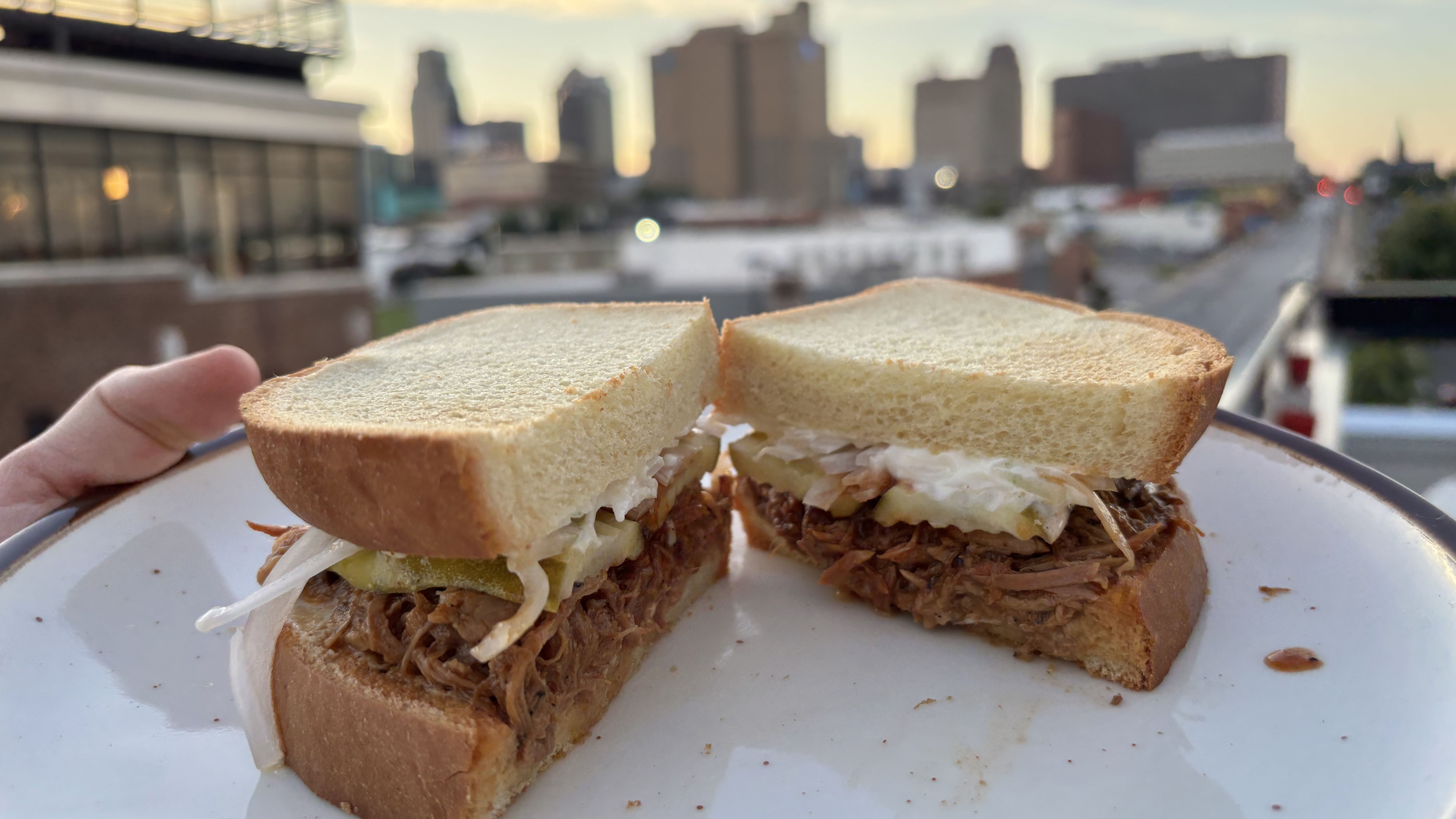 A pulled-pork sandwich on white bread, filled with slaw and pickles, cut in half on a plate. The blurred city skyline at sunset forms the urban backdrop.
