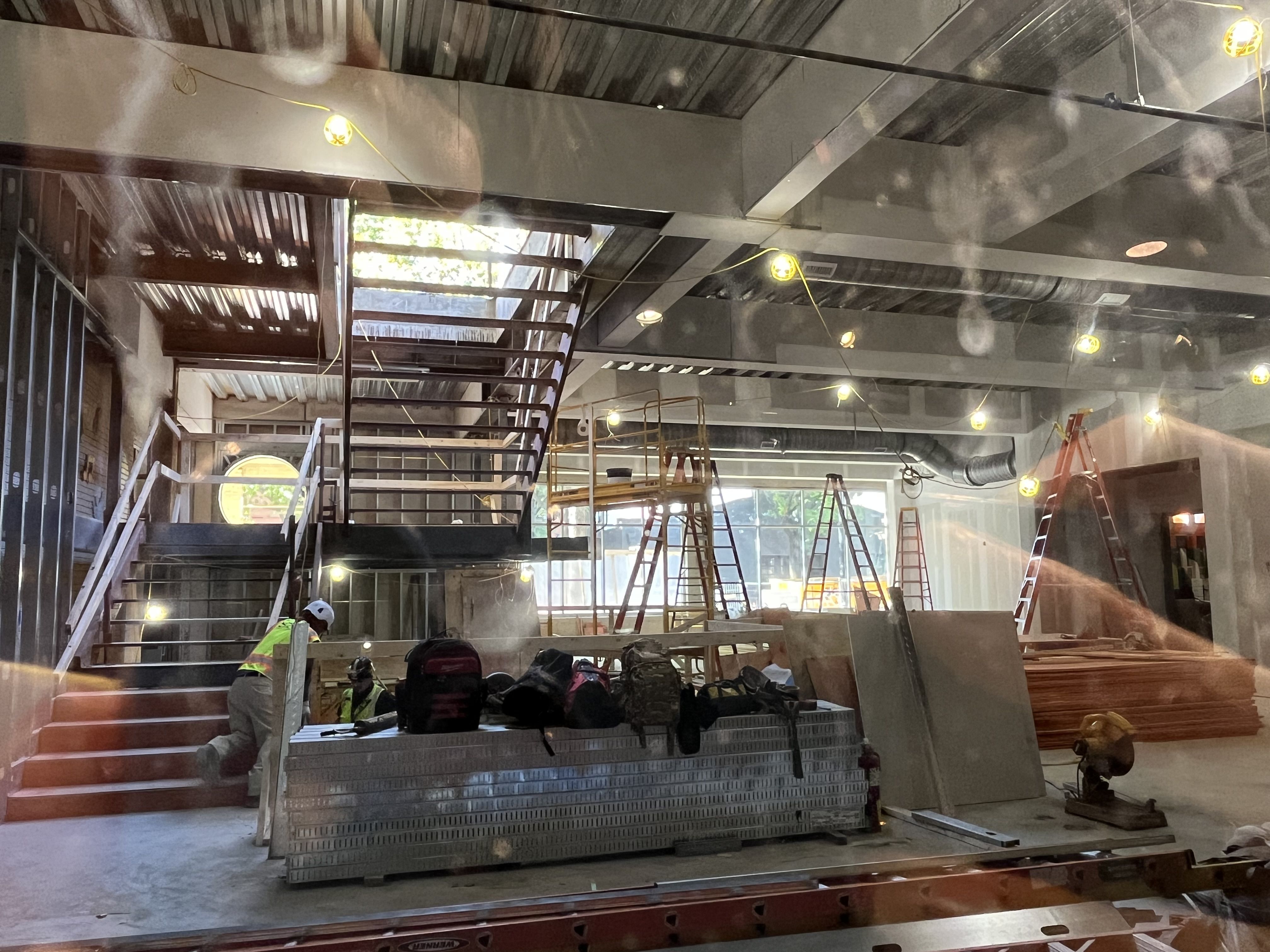 Construction site inside an unfinished building with exposed ducts and beams, metal stairs and scaffolding, and stacks of metal studs. Workers in hard hats and hi-vis vests beneath hanging yellow lights.