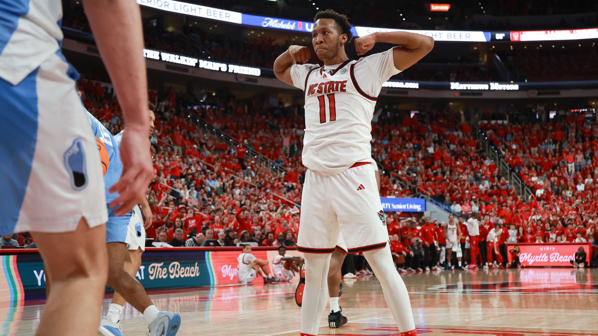 NC State basketball player in white uniform flexing arms in celebration on court, opposing players in light blue uniforms nearby, crowd in red shirts in background.