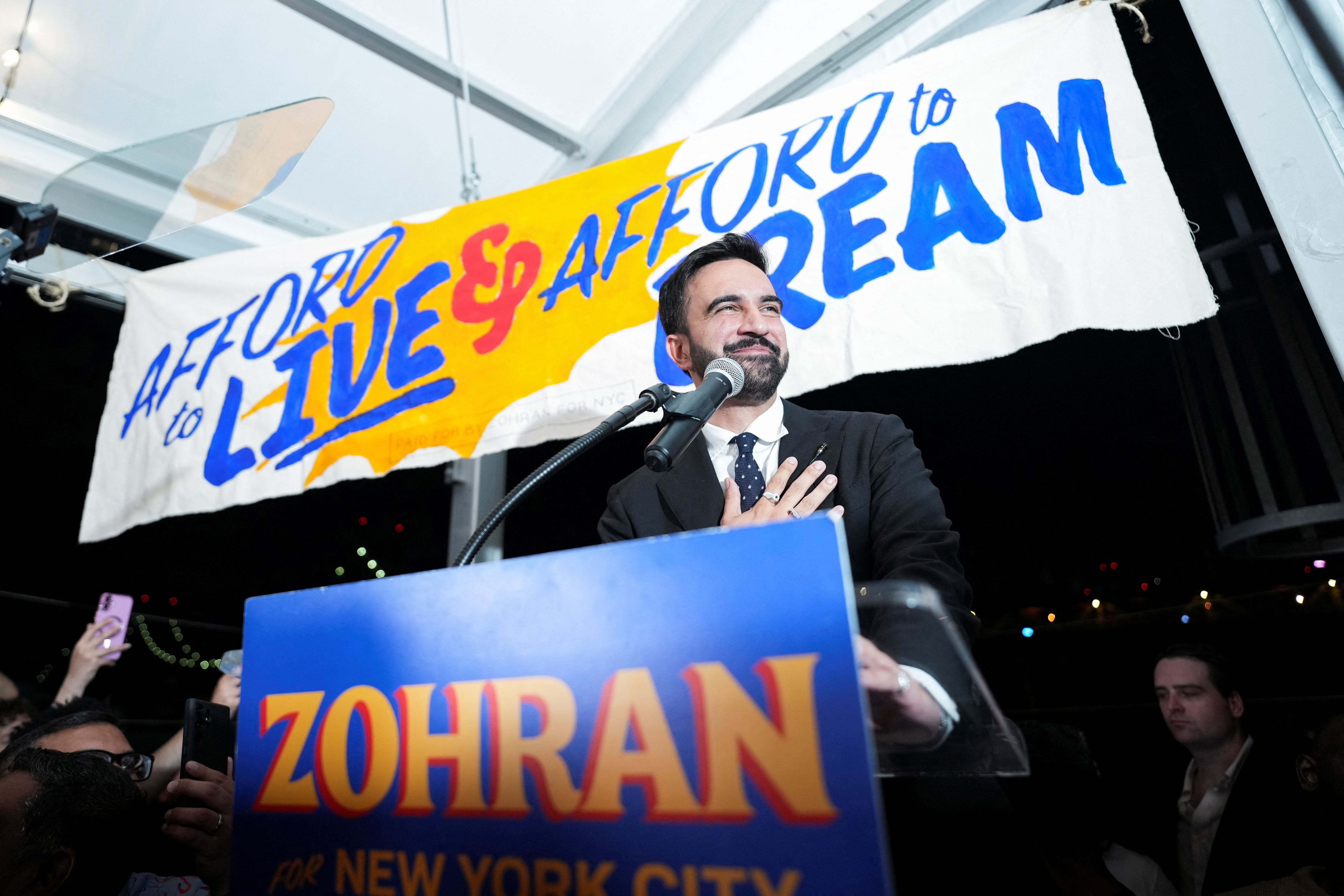 Zohran Mamdani. Behind him, a banner reads 'Afford to Live & Afford to Dream' in blue and red letters. The podium has a sign with the name 'Zohran' for New York City.