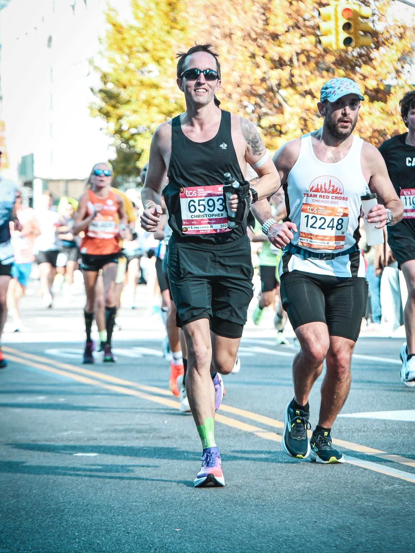 City street marathon in autumn. Front two runners: left, man in black tank top, sunglasses, holding a water bottle and green socks, bib 6593; right, man in white tank with cap, bib 12248.