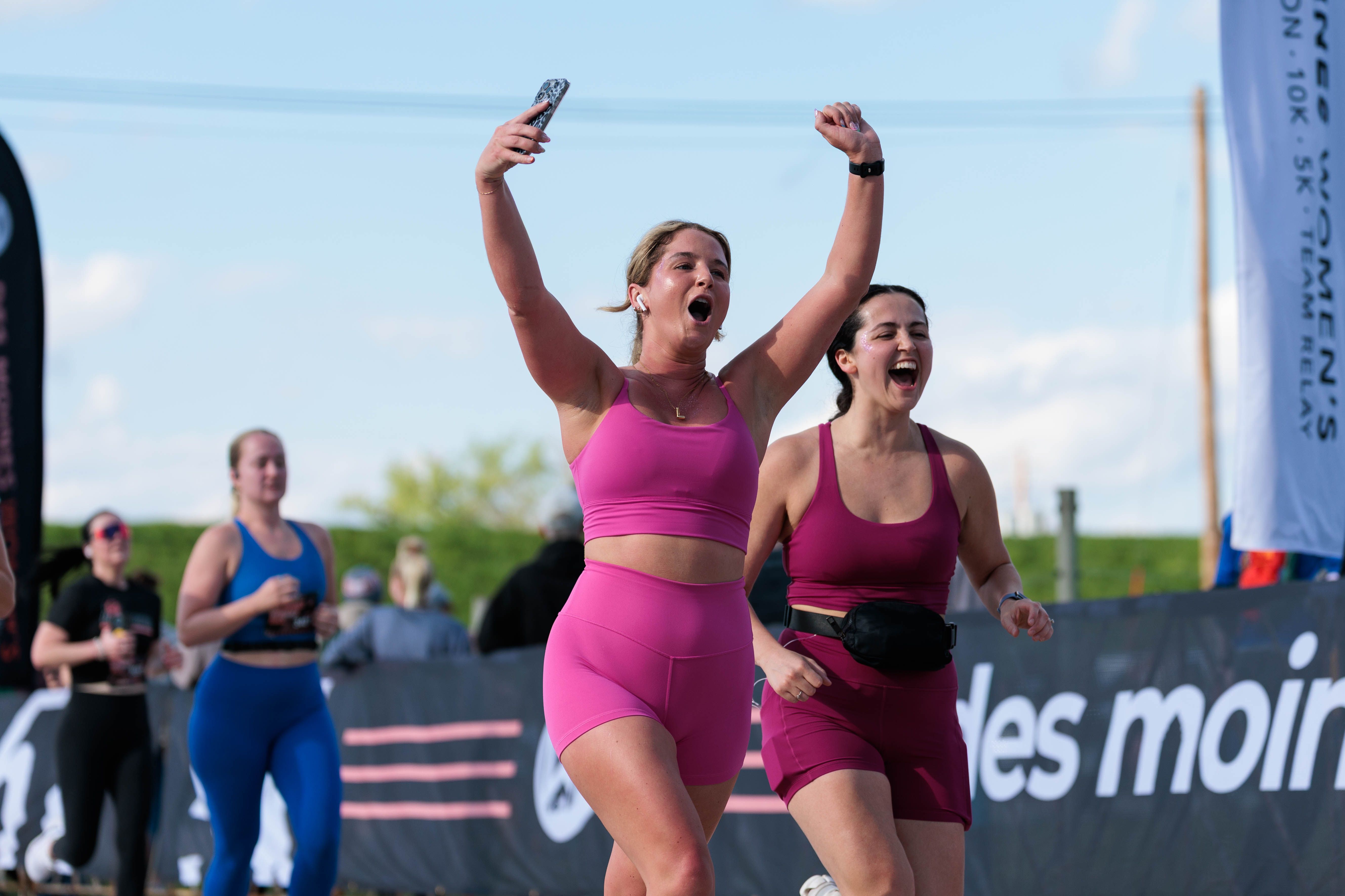 Two women in pink athletic outfits celebrate finishing a race, arms raised in triumph, with spectators and event banners along the course under a blue sky.
