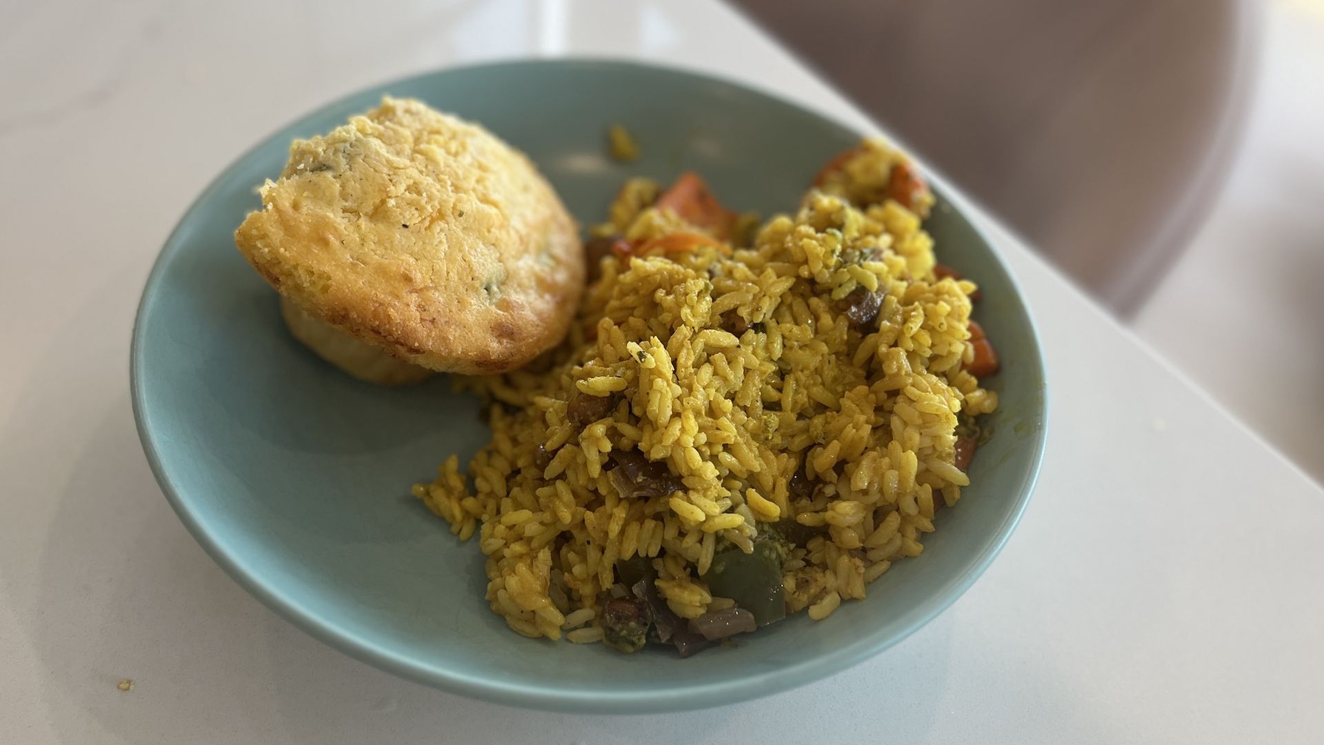 Blue plate on a pale table holds yellow rice with vegetables and two golden-brown biscuits; a blurred arm is visible in the background.