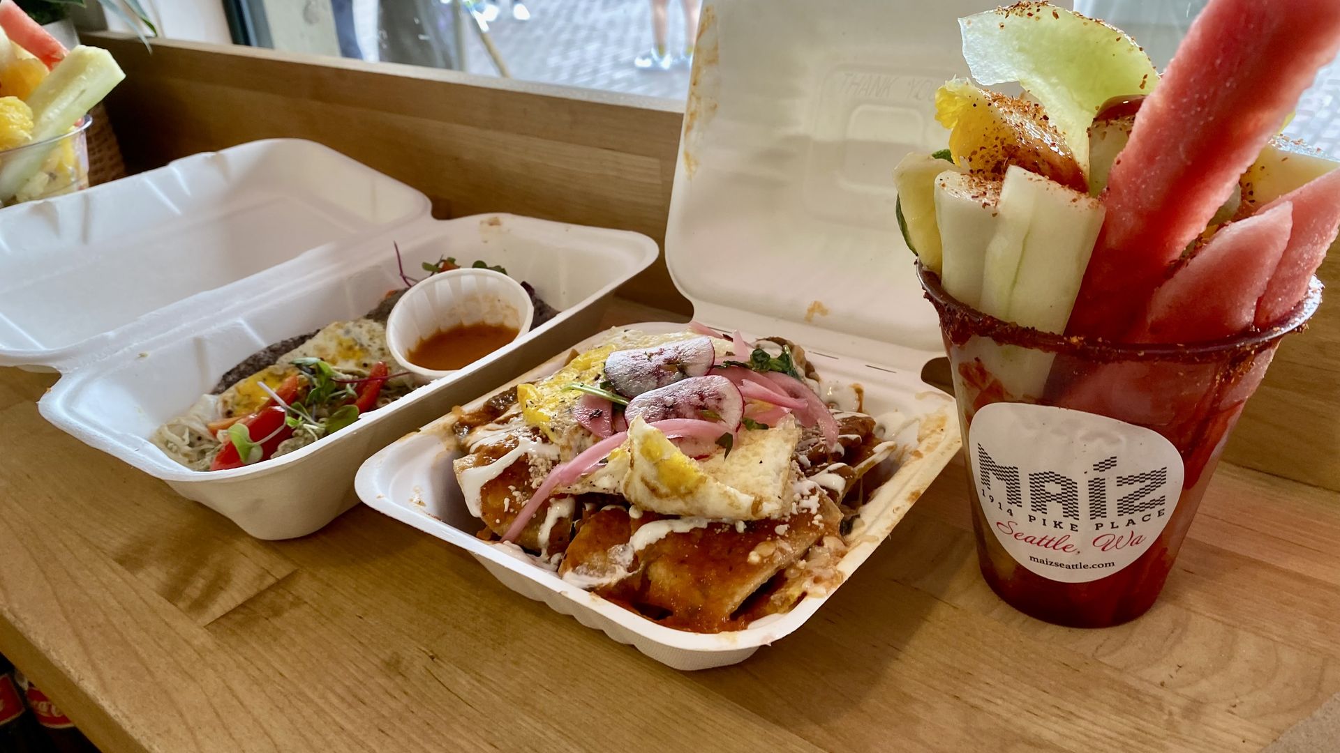 Two two go containers, one with tacos and the other with chilaquiles, next to a cup filled with spears of cut fruit.