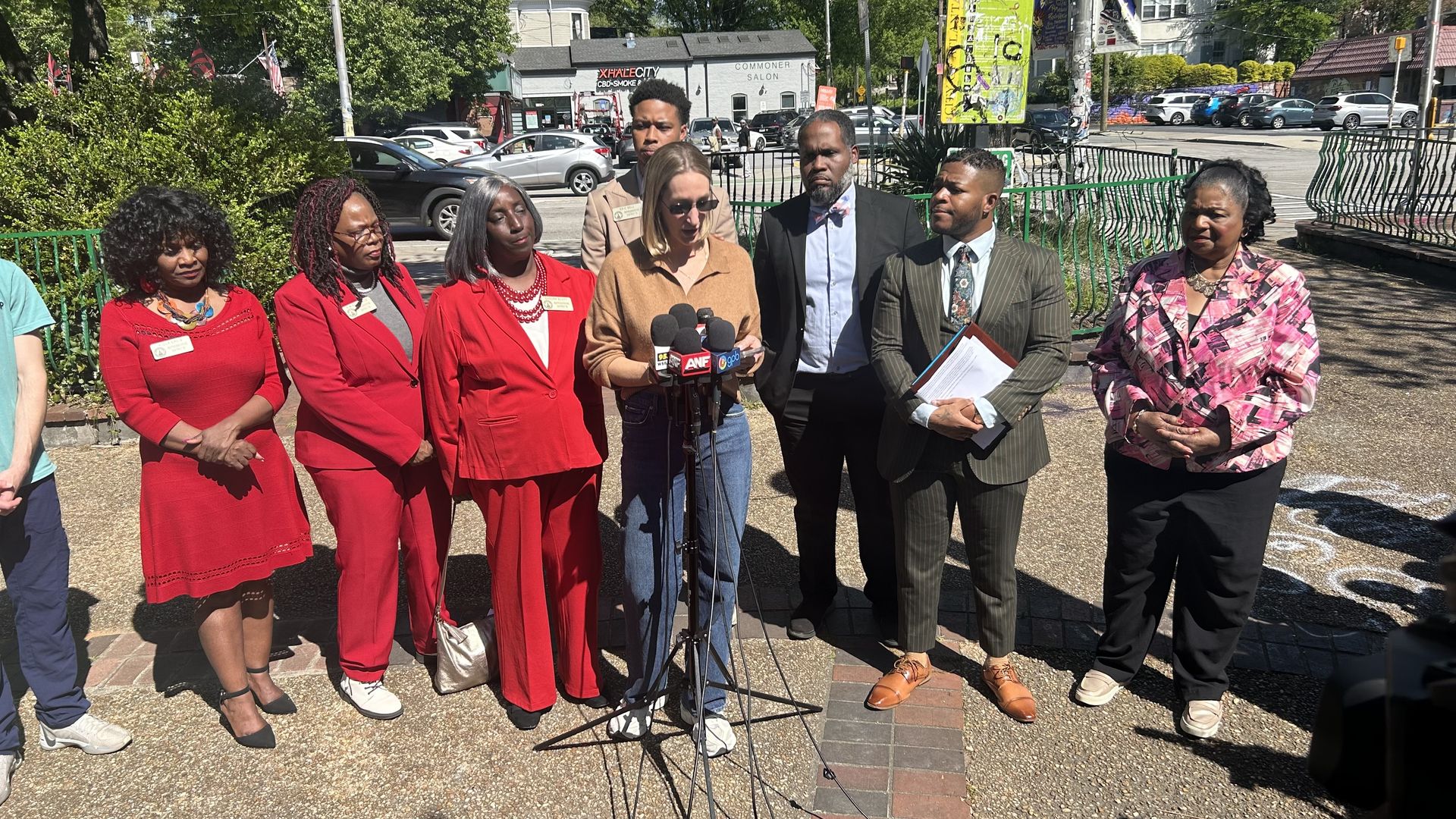 A group of people stand in an urban park along a busy street and hold a press conference