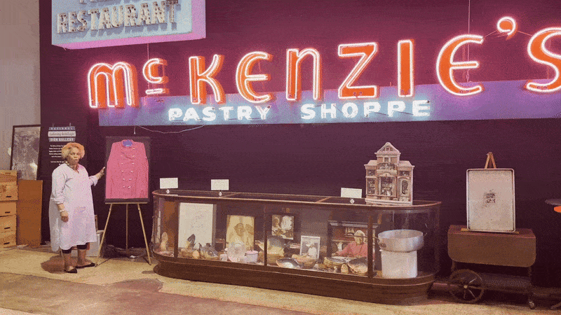 A woman gestures toward a pink chef's coat underneath a neon sign for McKenzie's Pastry Shoppe.