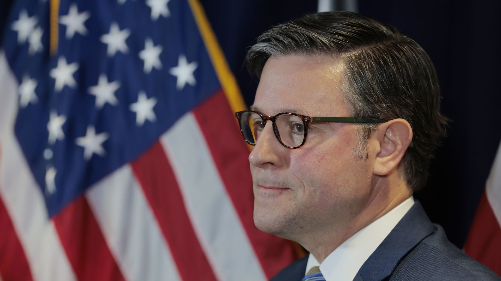 Mike Johnson, wearing a blue suit and glasses and standing in front of American flags.