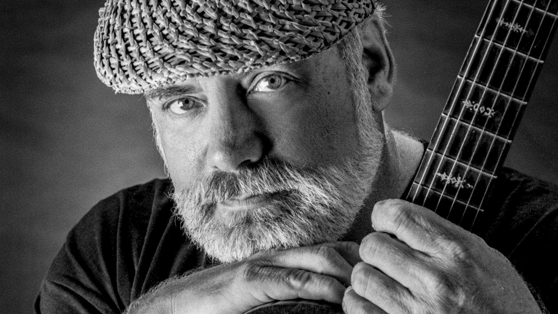 Black and white close-up portrait of a bearded man wearing a woven hat, holding and leaning on an acoustic guitar, with a calm, thoughtful expression.