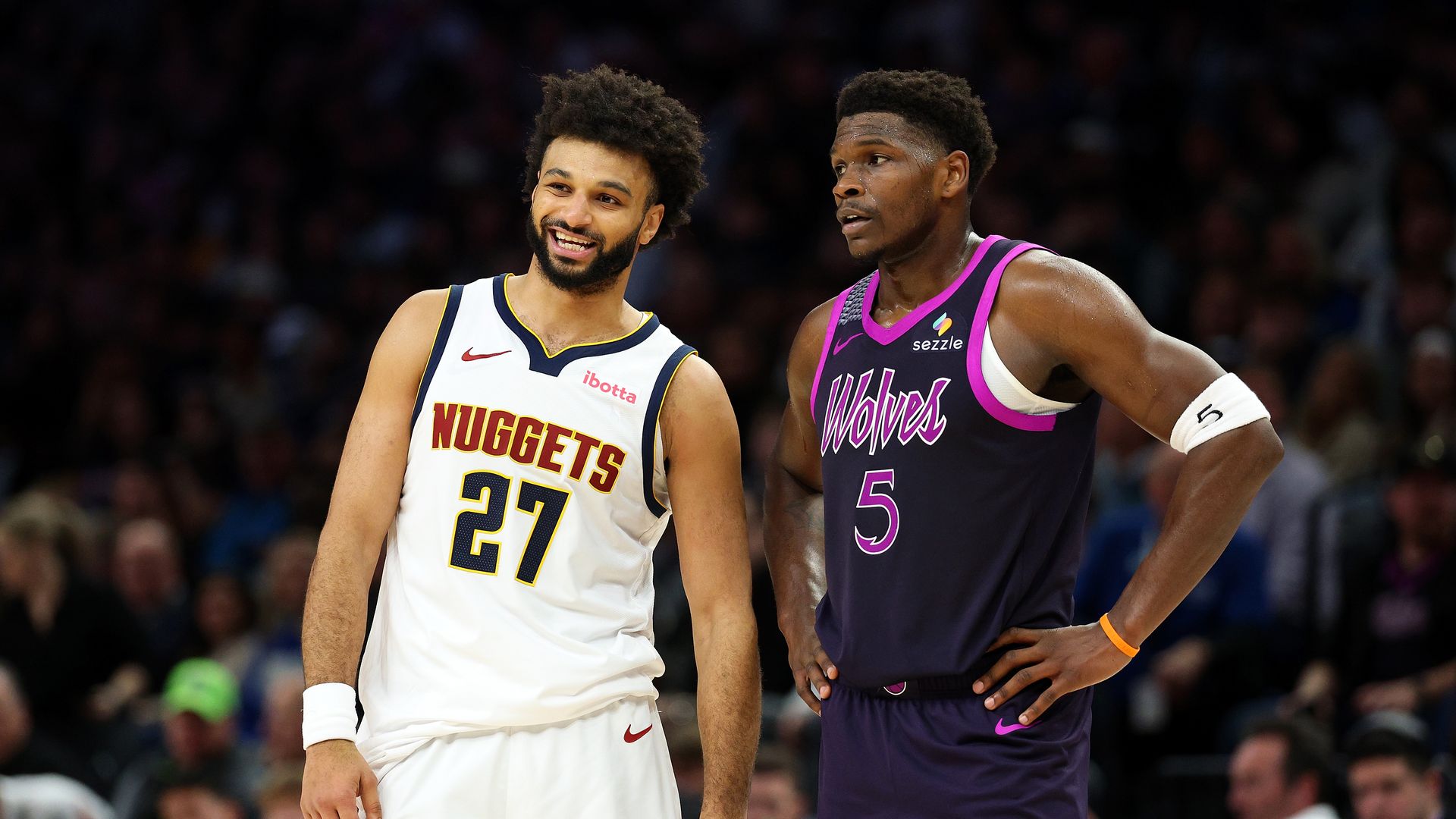 Two basketball players stand on a court; Jamal Murray in white jersey smiles beside Anthony Edwards in purple, as a blurred crowd watches.