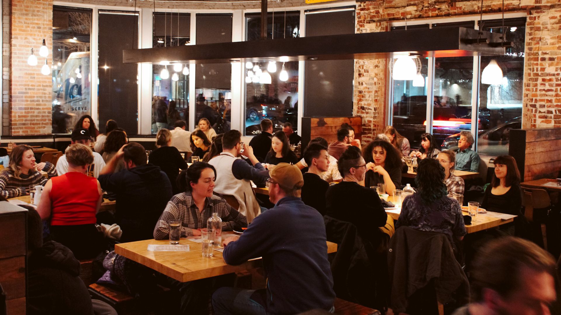 Busy restaurant scene with exposed brick walls, hanging pendant lights, and long wooden tables crowded with diners; a colorful sign on the brick wall reads Mixed Drinks.