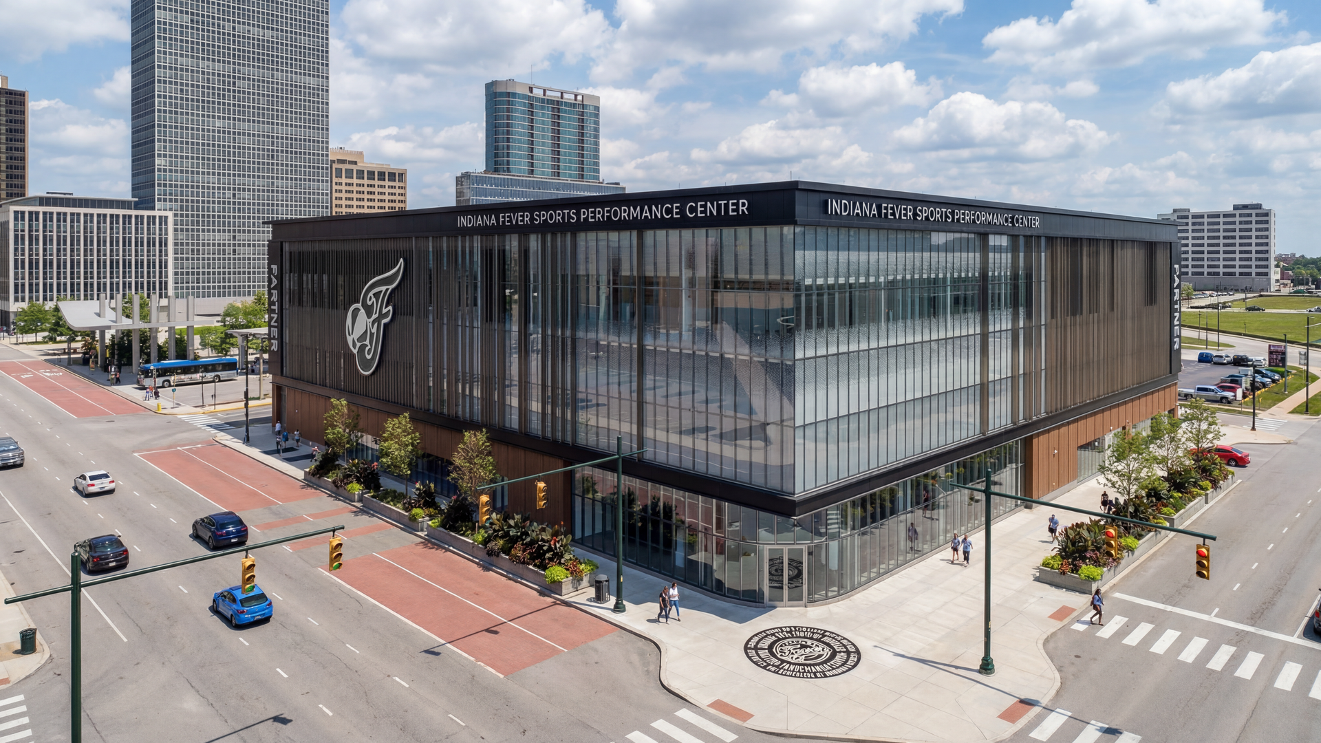Modern glass-front building labeled "Indiana Fever Sports Performance Center" at a busy intersection, with a large side logo, pedestrians on the sidewalk, and tall city buildings under a blue sky.