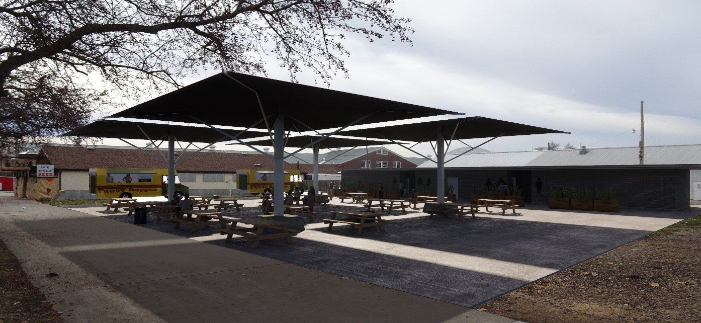 Outdoor seating area with multiple wooden picnic tables under large black shade canopies, adjacent to a gray building and two yellow food trucks in the background on a cloudy day.