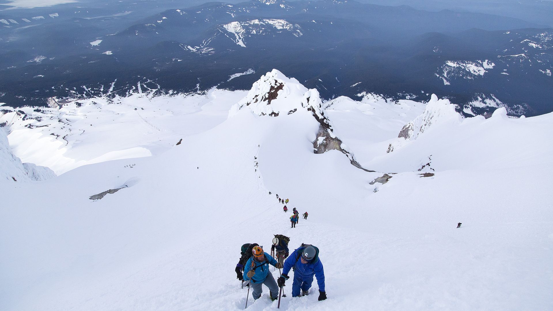 Group of climbers in colorful winter gear ascending a snowy mountain slope, with vast snow-covered peaks and dark forested hills in the background under a clear sky.