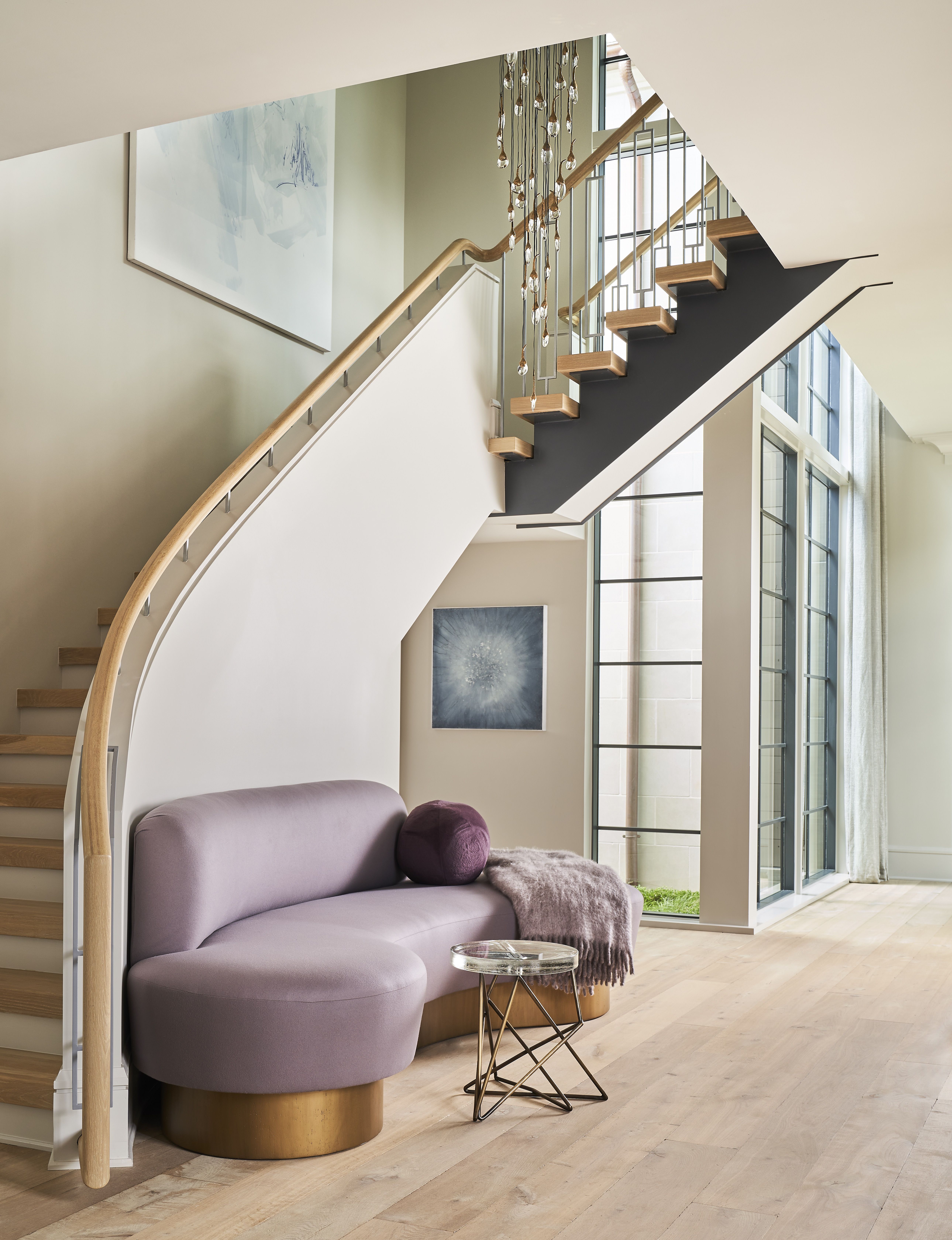 Modern interior with a diagonal floating wooden staircase and black metal stringer, crystal chandelier strands, pale lavender curved sofa, a glass side table, large windows, and abstract blue wall art.