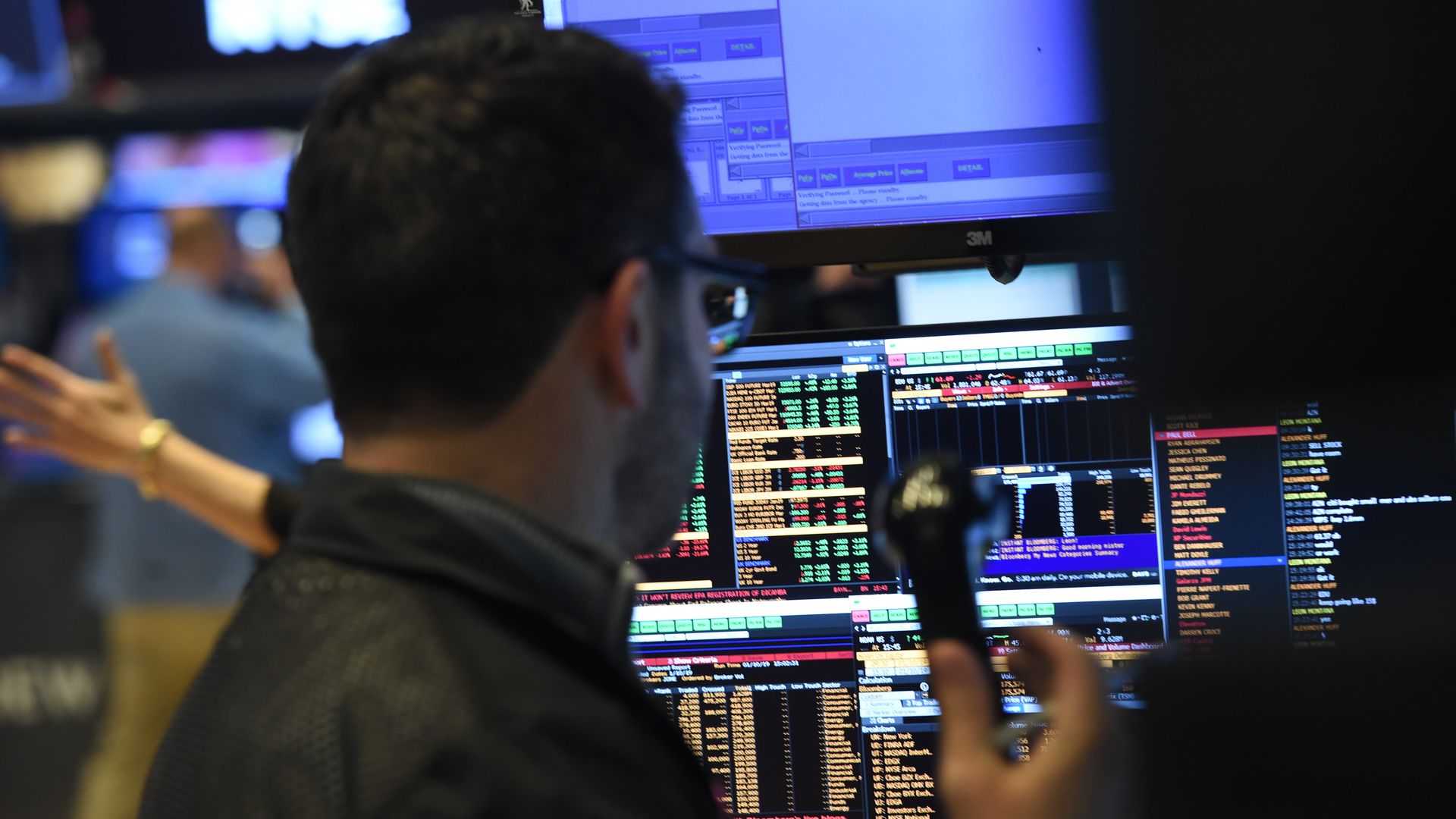 In this image, a broker at the New York Stock Exchange stands with a telephone in front of screens. 