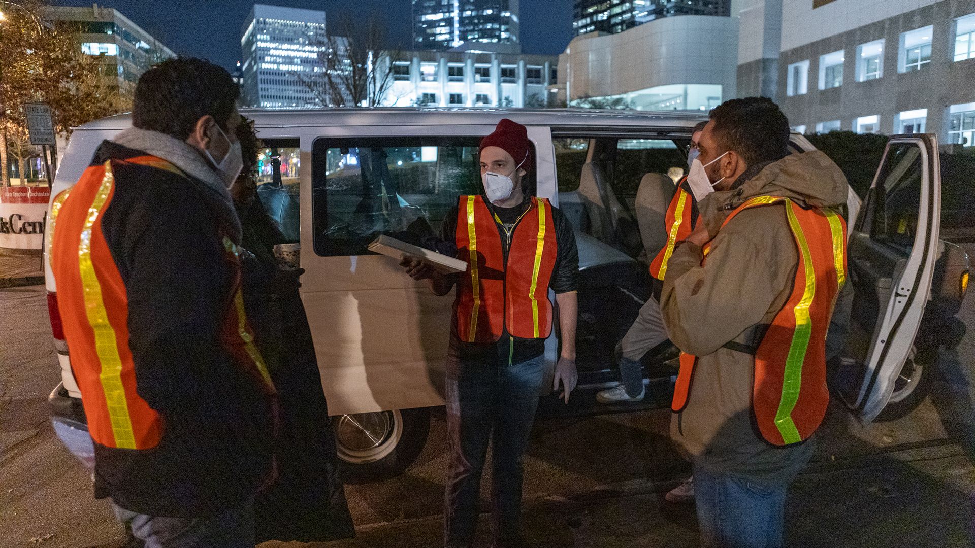Several people wearing Covid masks and reflective vests stand in front of a white van at night before conducting a census of homeless people
